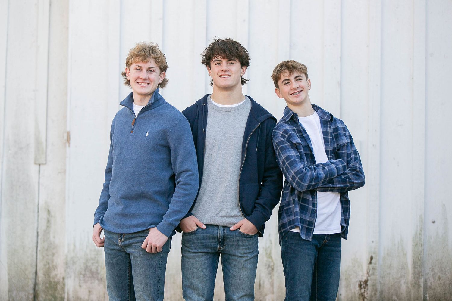 Three teenagers standing against a white wall wearing casual clothing in blue and grey tones.