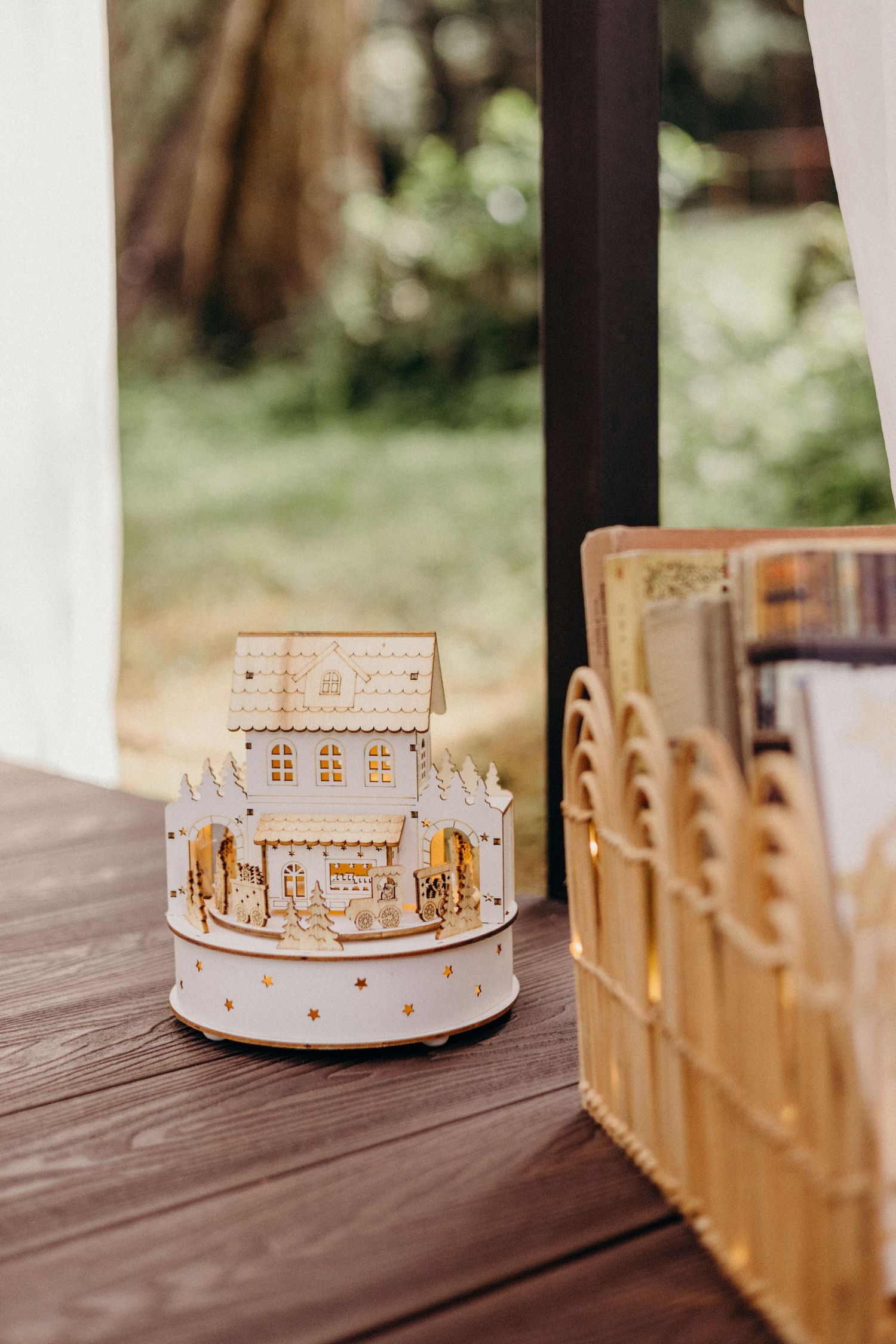 A white decorative light-up house music box sits on a wooden table with soft bokeh background.