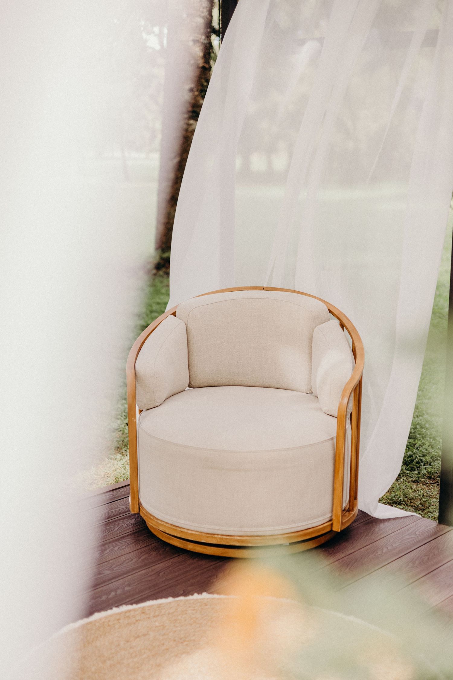 A modern curved beige upholstered armchair with wooden frame sits elegantly on a wooden floor near white curtains.