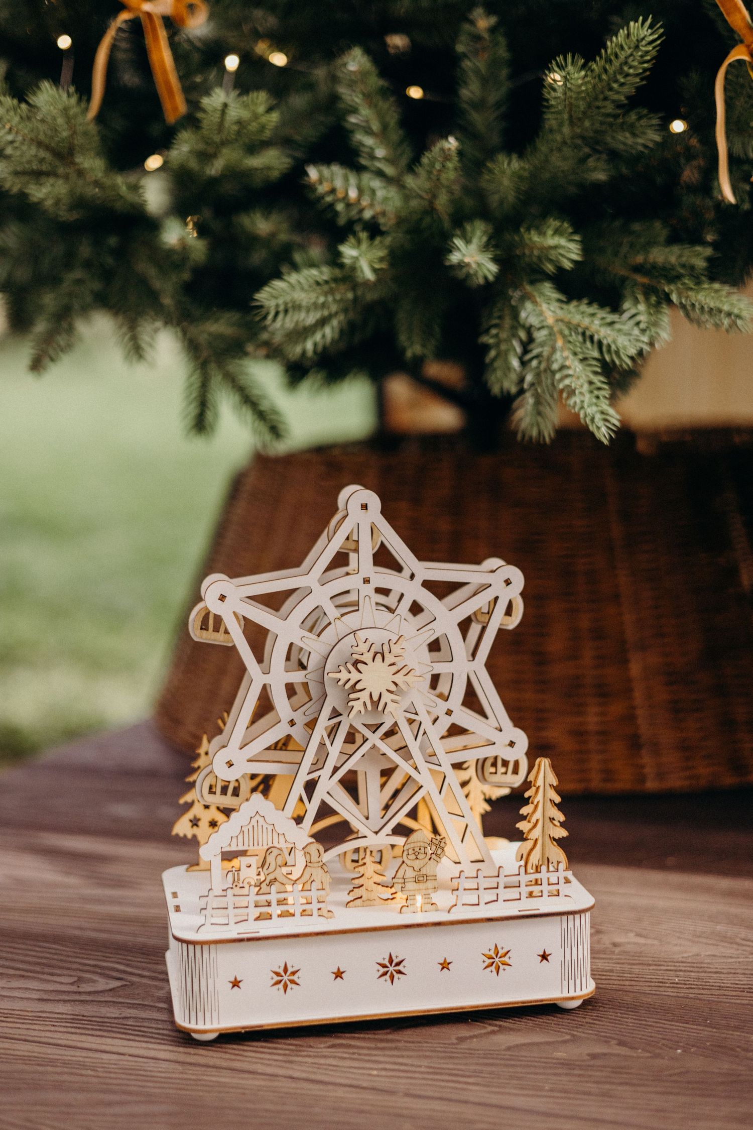 White wooden Ferris wheel Christmas music box decoration with gold accents sits beneath evergreen branches on wooden surface.