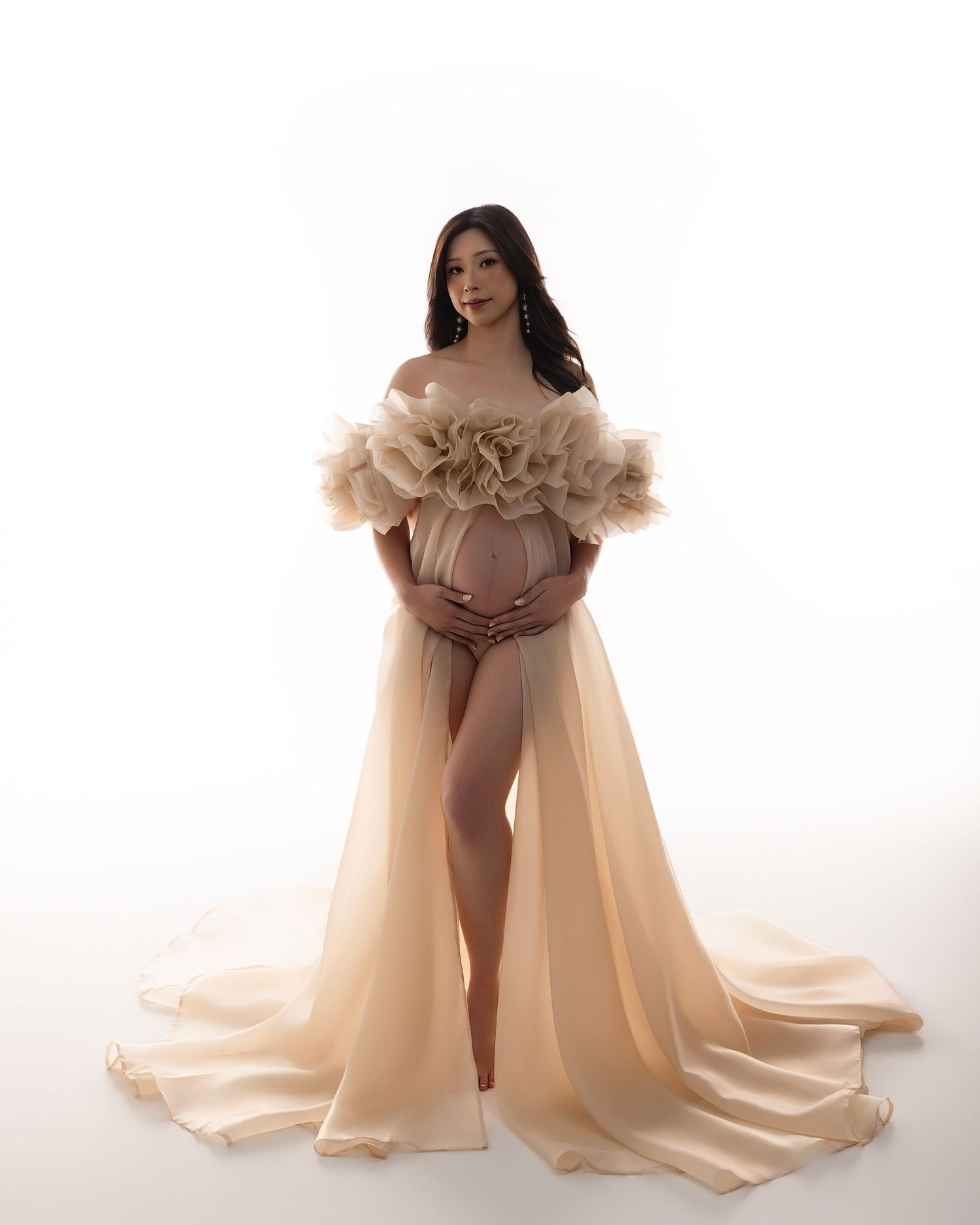 Expectant mother posing in sheer champagne tulle maternity dress with ruffled bodice and long flowing skirt.