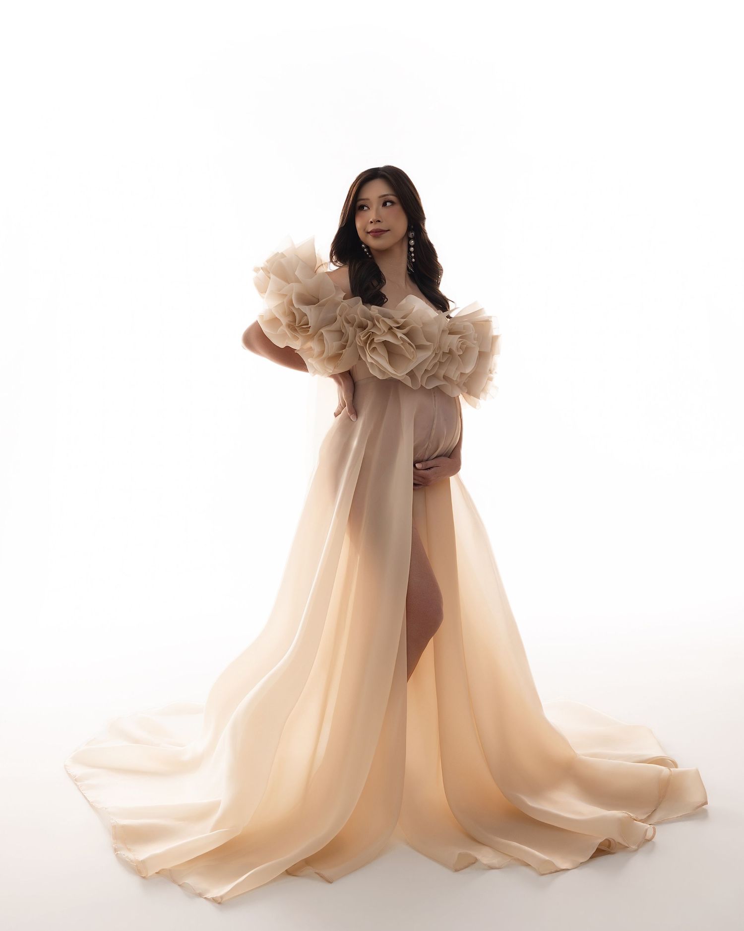 Pregnant woman in elegant champagne ruffled off-shoulder maternity gown with flowing train, studio photo.