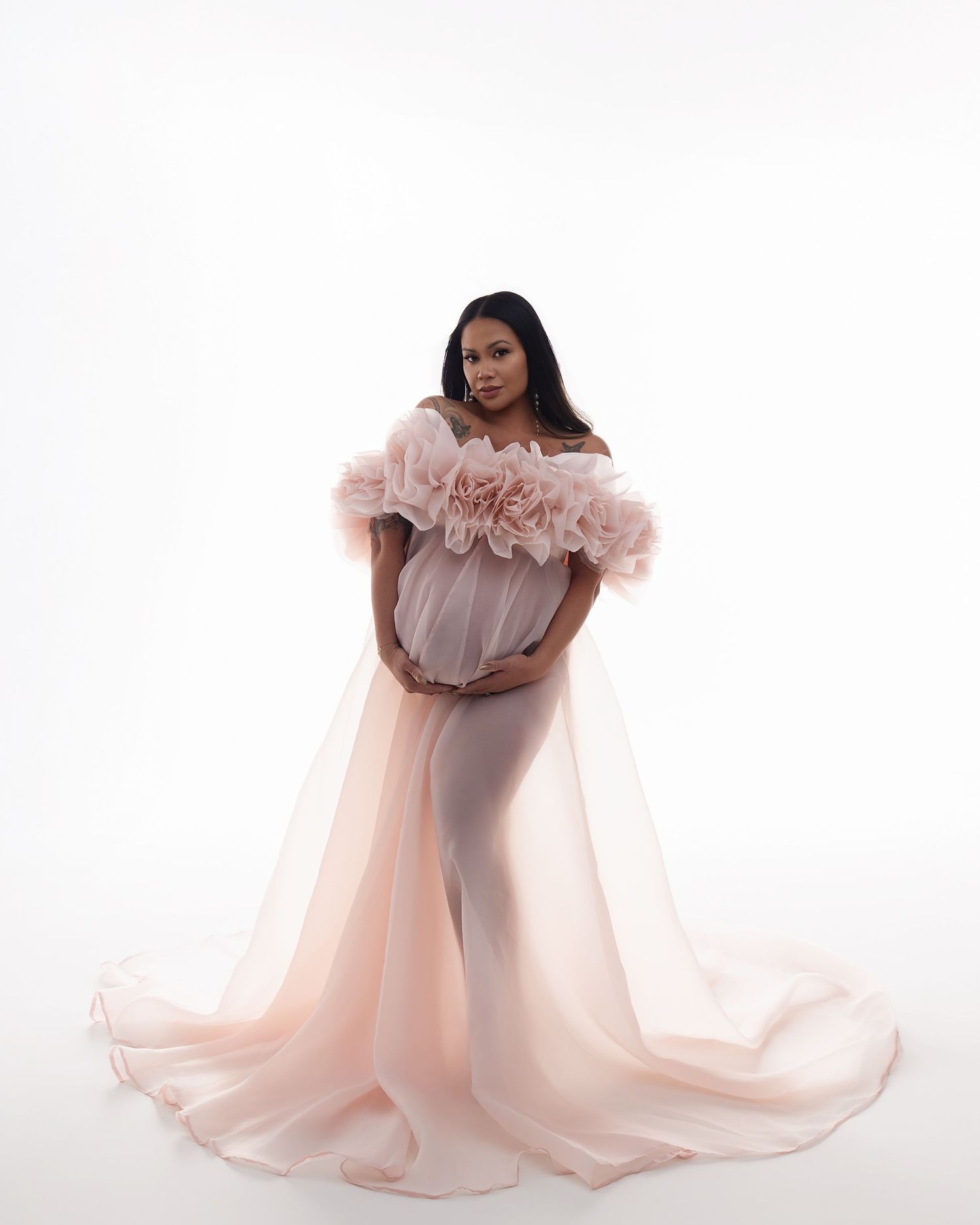Pregnant woman in blush pink off-shoulder tulle maternity gown with floral details and flowing train on white background.