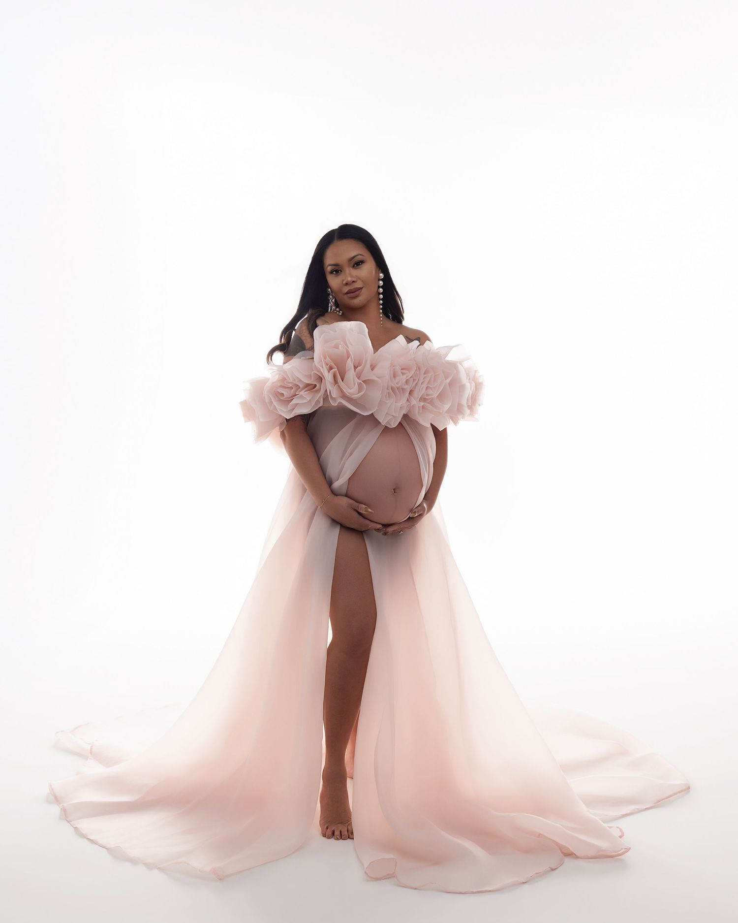 Pregnant woman in elegant blush pink ruffled tulle maternity gown posing gracefully against white background.