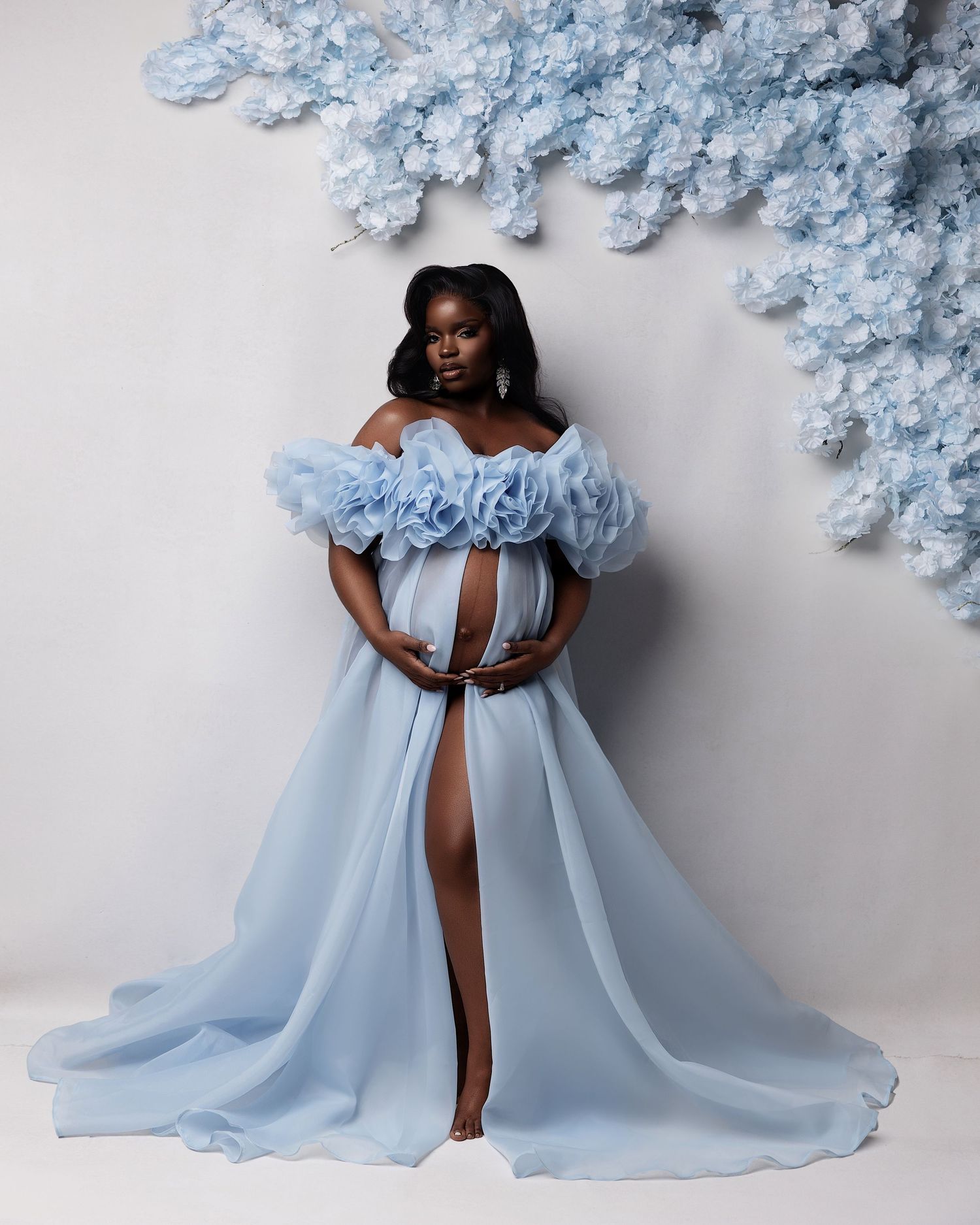 Pregnant woman in elegant light blue off-shoulder ruffle gown with floral backdrop for maternity photoshoot.