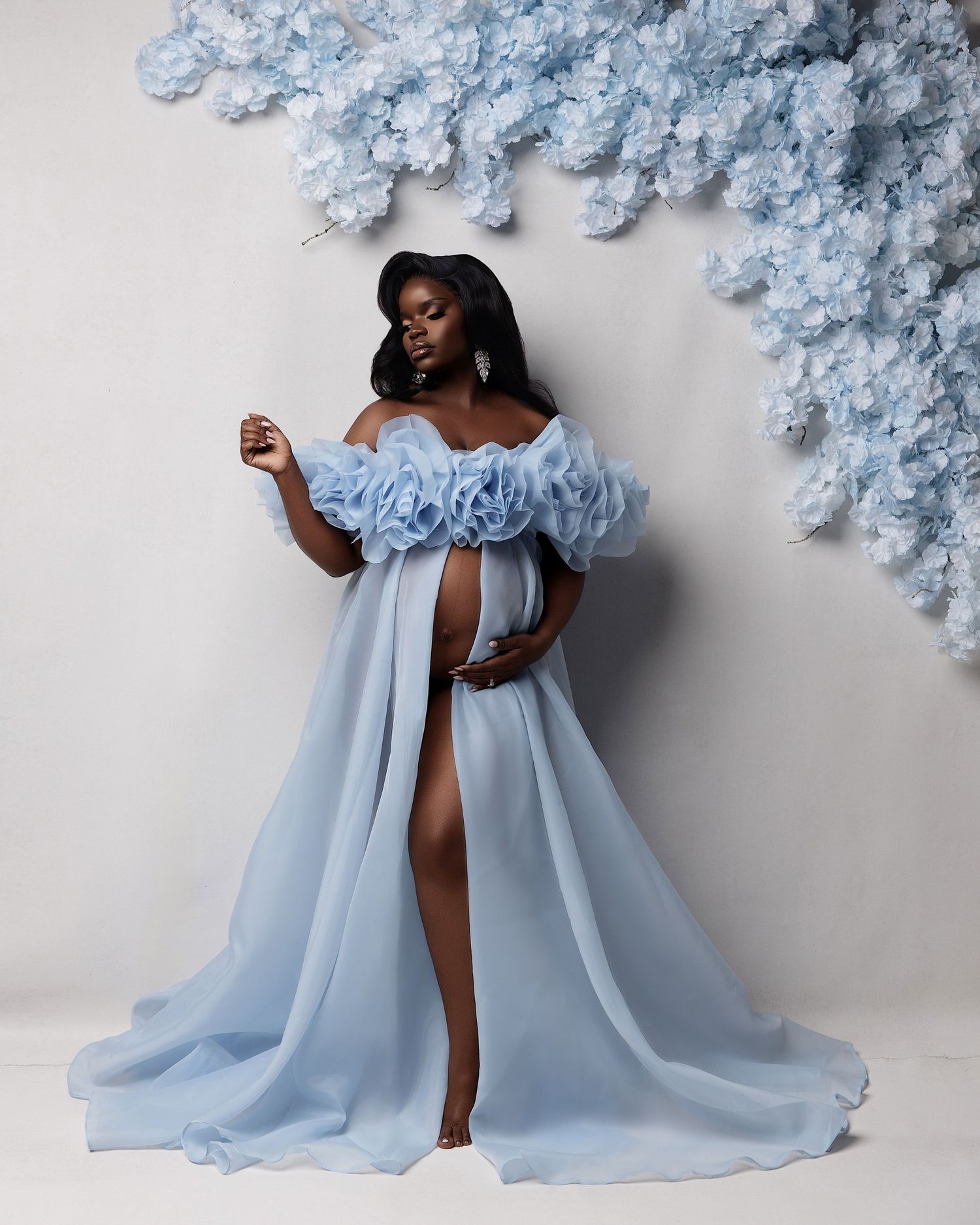 Pregnant woman in elegant light blue ruffled off-shoulder maternity gown with floral backdrop for a professional photoshoot.