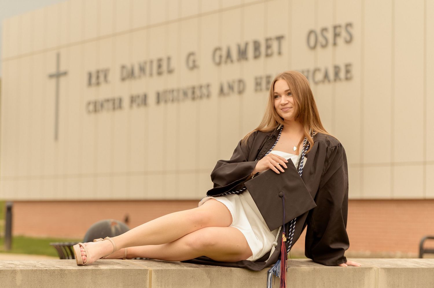A graduate in cap and gown poses by the York College entrance sign on a spring day.