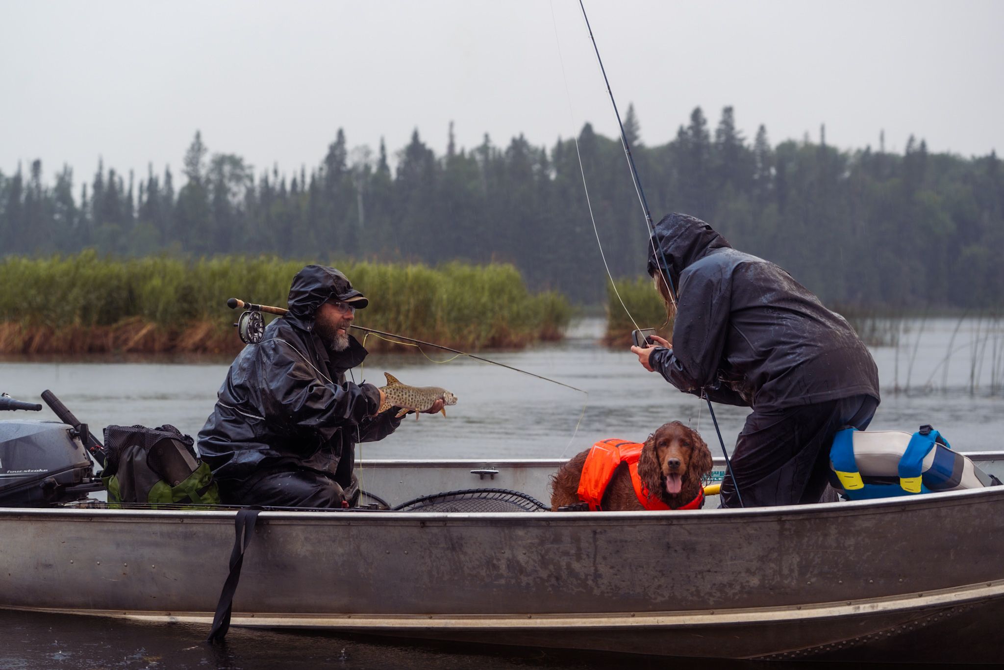 Two anglers in rain gear fish from an aluminum boat on a cloudy day with evergreen trees in the background.
