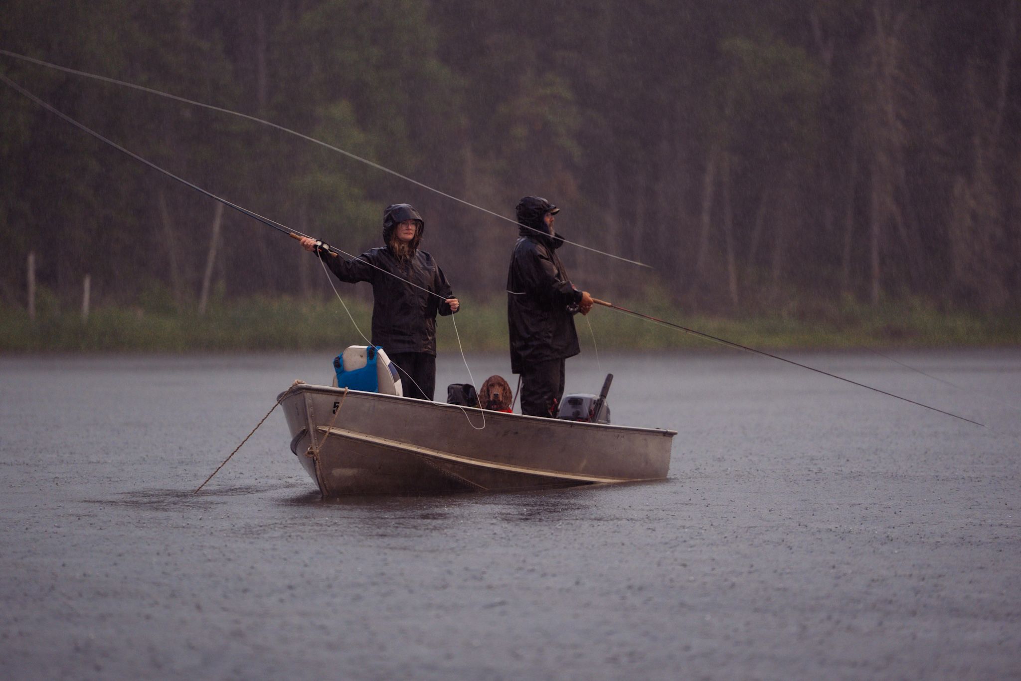 Two people fishing from a small boat in the rain on a misty lake with fishing poles extended.