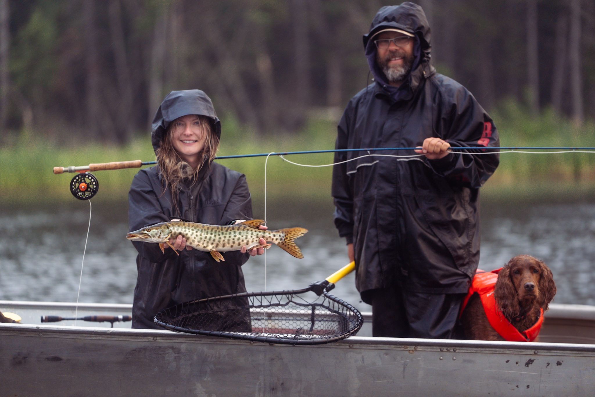 Two people wearing rain gear fish from a boat in wet weather, displaying a caught pike while a dog watches.