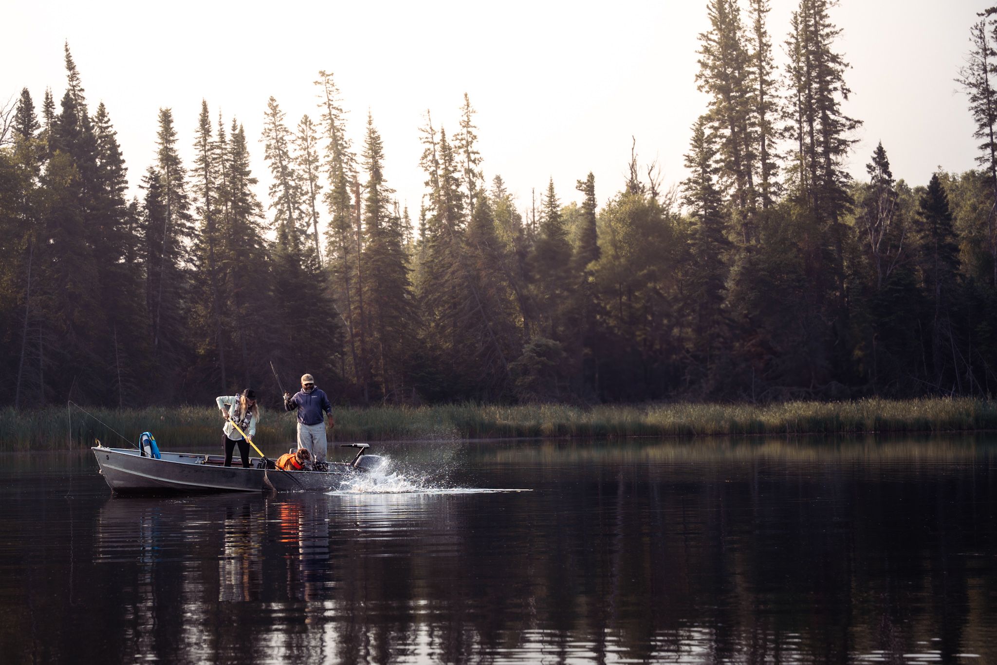 People fishing from a canoe at sunset on a tranquil lake surrounded by evergreen trees.