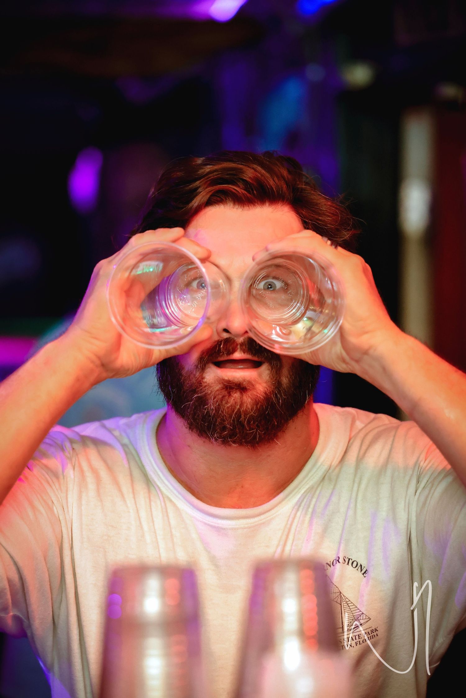 A bearded person makes binocular shapes with their hands in front of their eyes at a party.