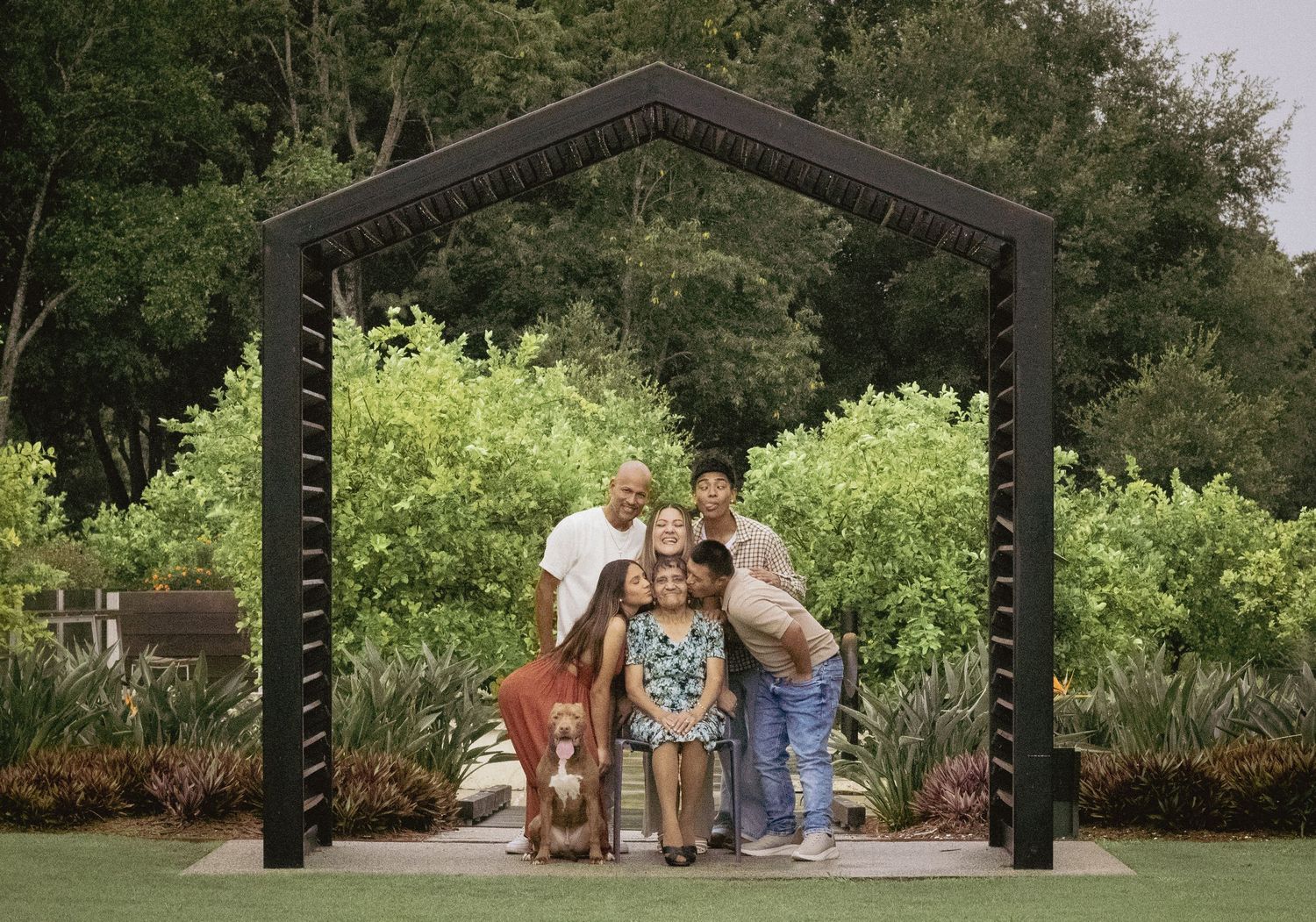 Multi-generational family portrait under decorative arch surrounded by lush green trees outdoors.