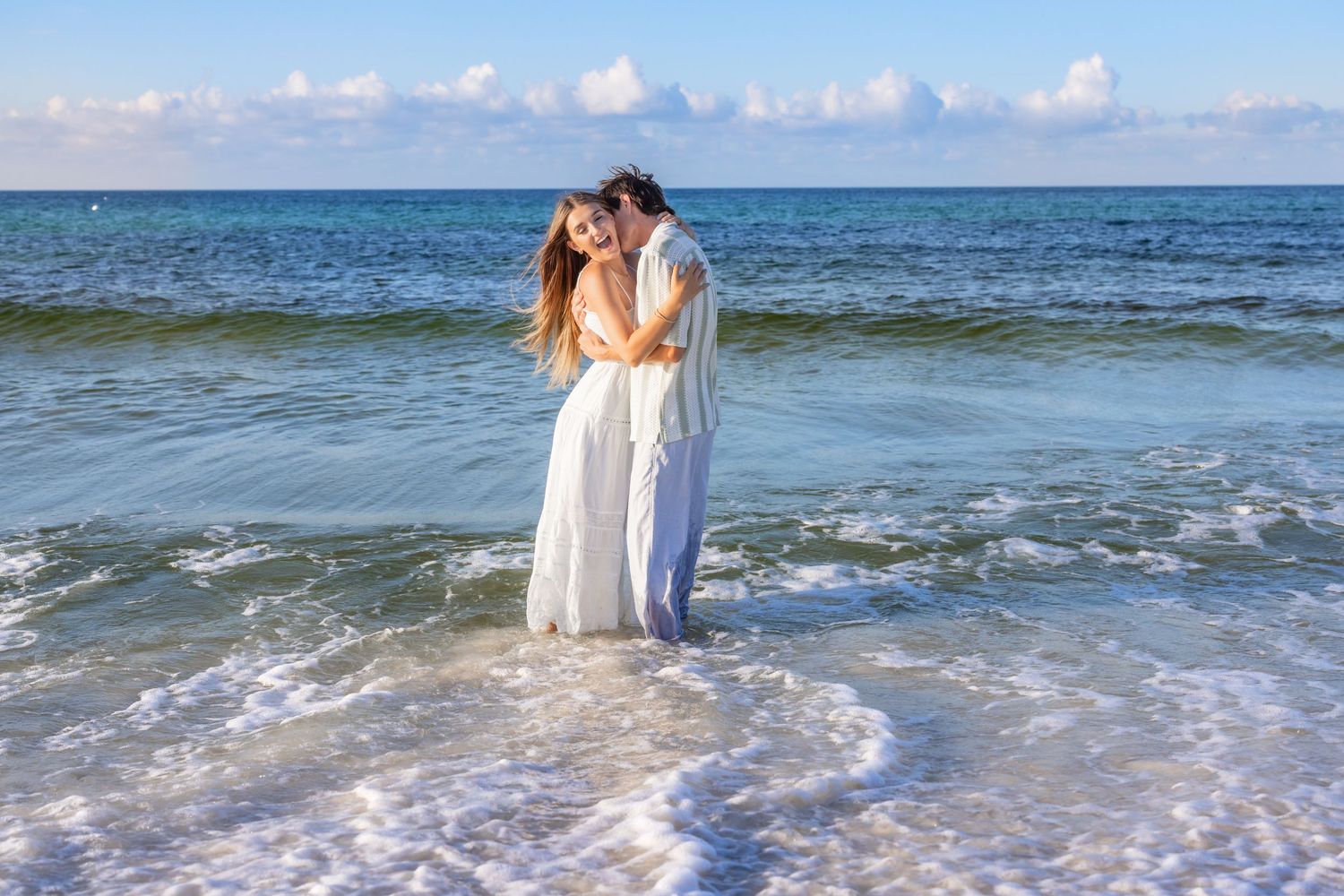 A romantic couple in white dresses embraces on a sunny beach with a pier visible in the distance over blue waters.