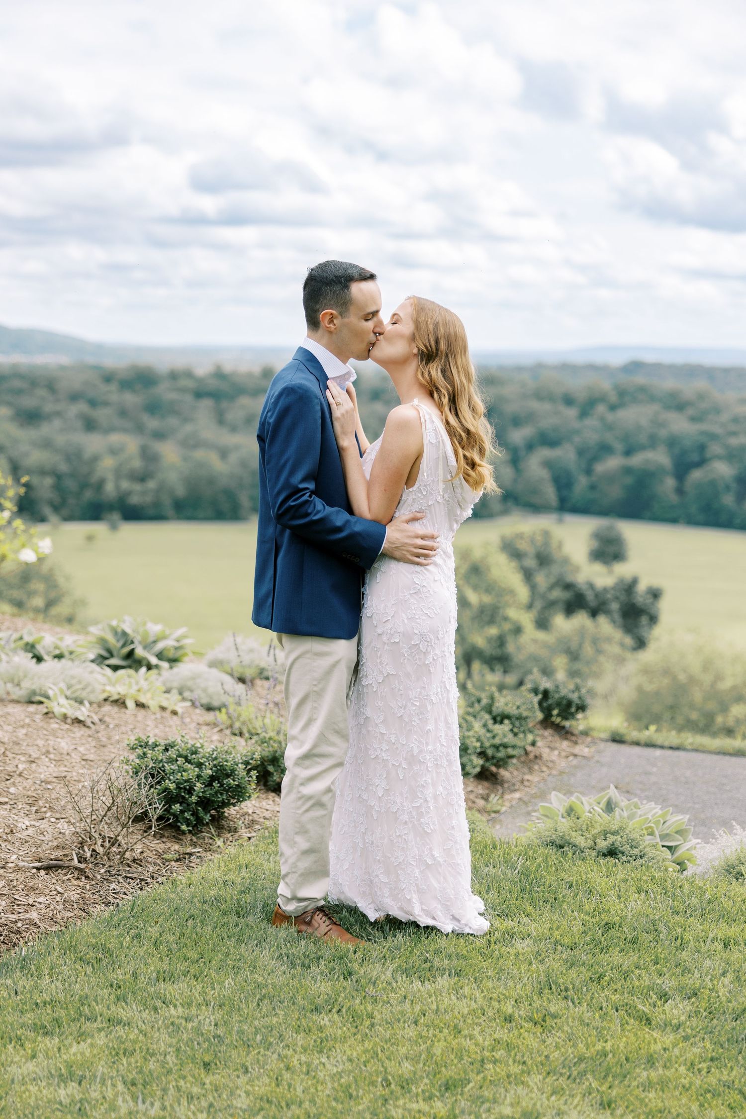 A couple in wedding attire shares an intimate moment on a hilltop overlooking a lush green landscape on their special day at Pendry Natirar.