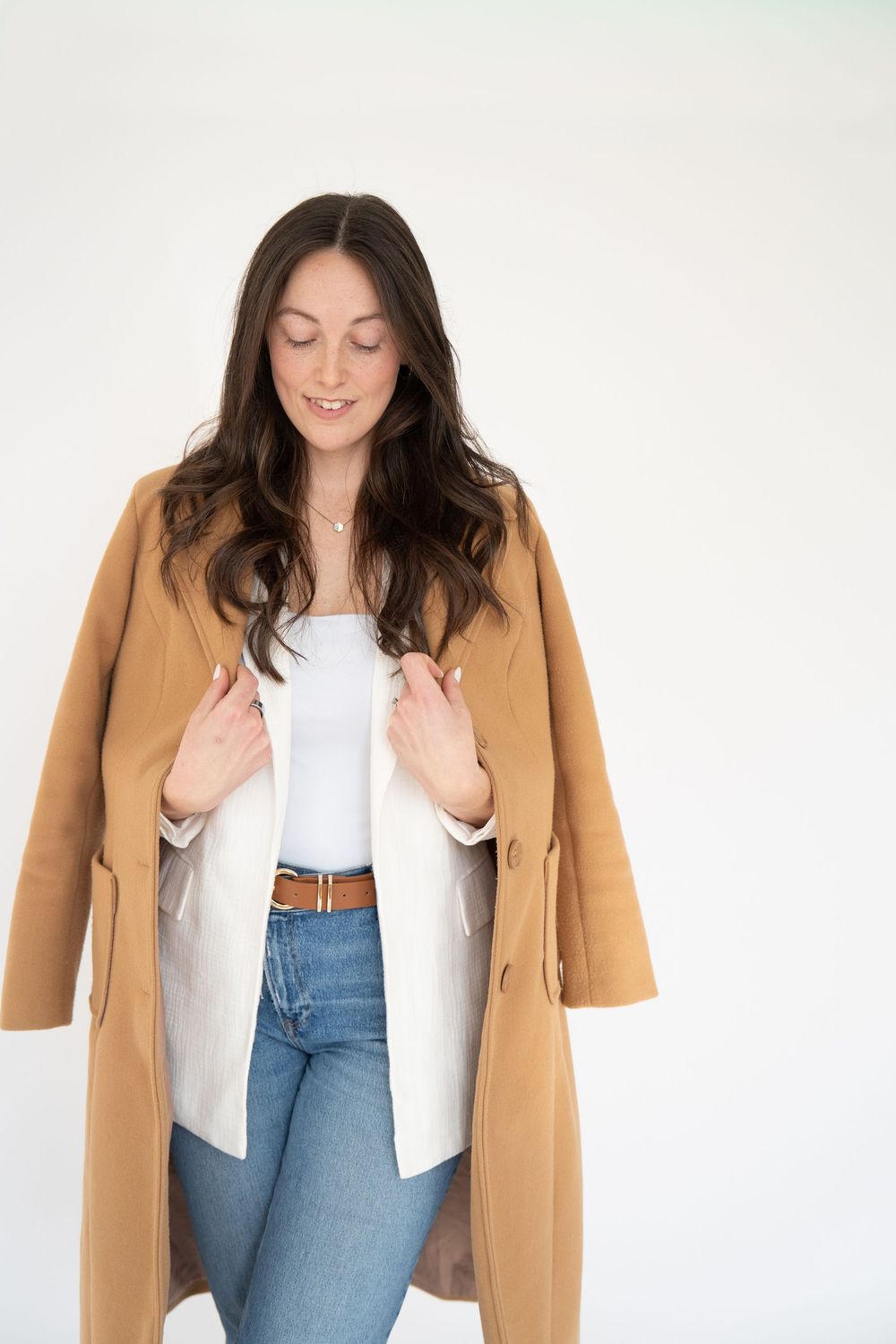 Woman in camel coat, white top, and jeans posing against white background.