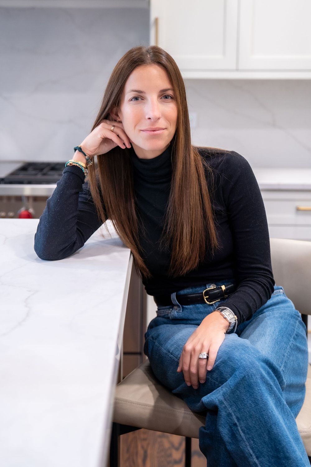 Woman in black turtleneck and jeans sits relaxed at a marble kitchen island, smiling naturally in a modern white kitchen.