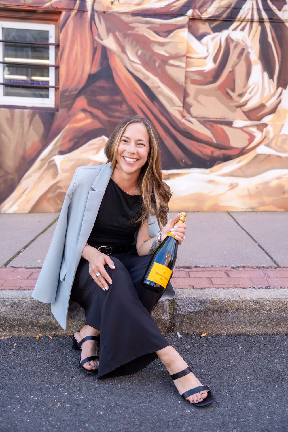 Smiling woman in black outfit and gray blazer sitting outdoors holding Veuve Clicquot champagne bottle by colorful mural.