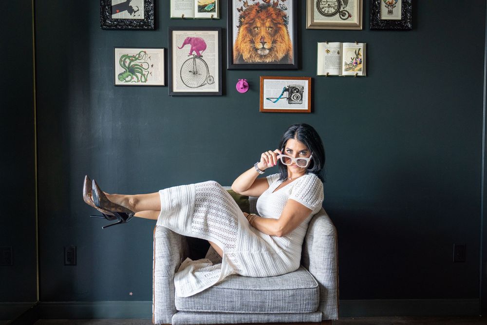 Woman relaxing in armchair with legs up, wearing white outfit and glasses, surrounded by eclectic art gallery wall.