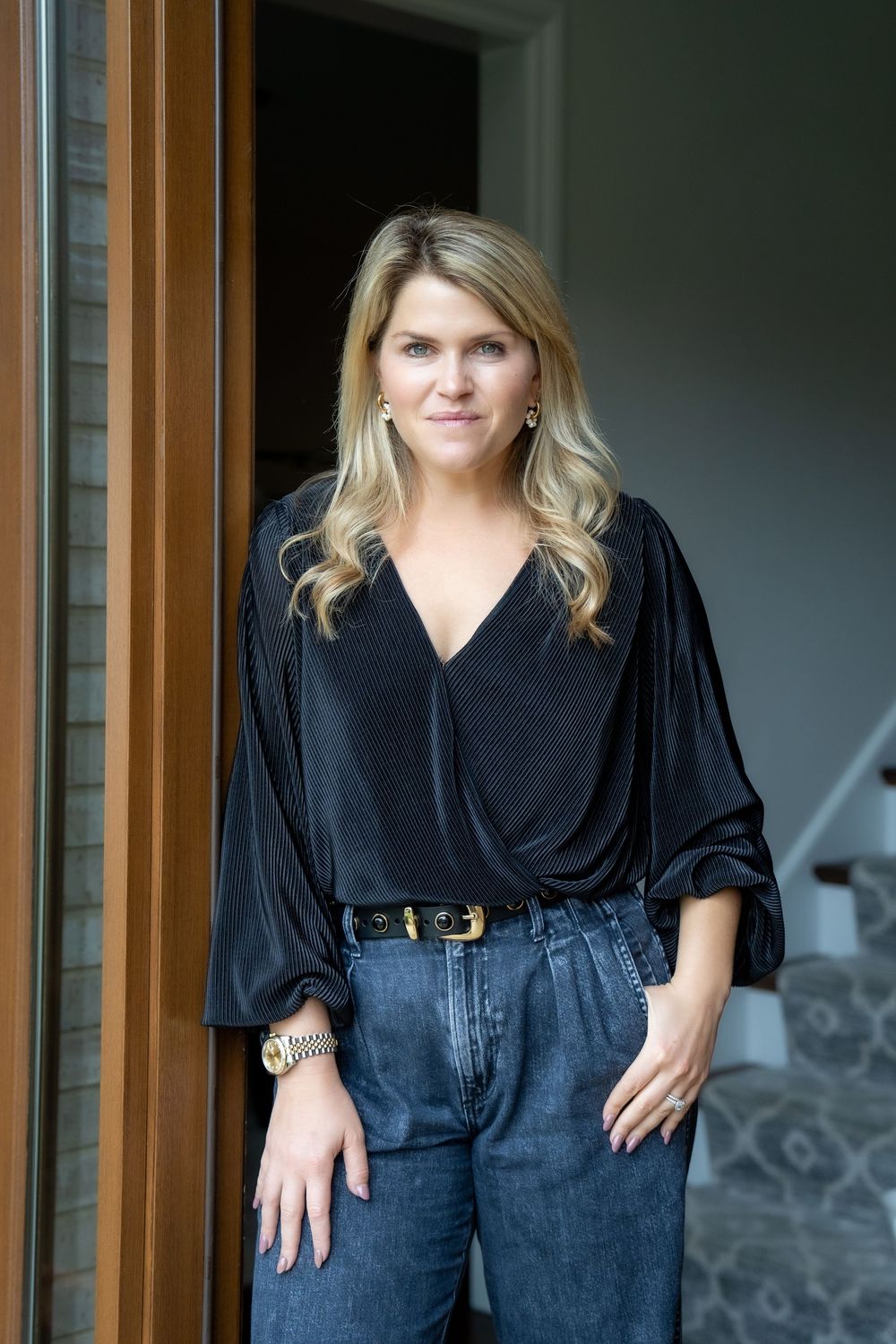 Blonde woman in black velvet blouse and jeans posing by a wooden door in a stylish home interior.