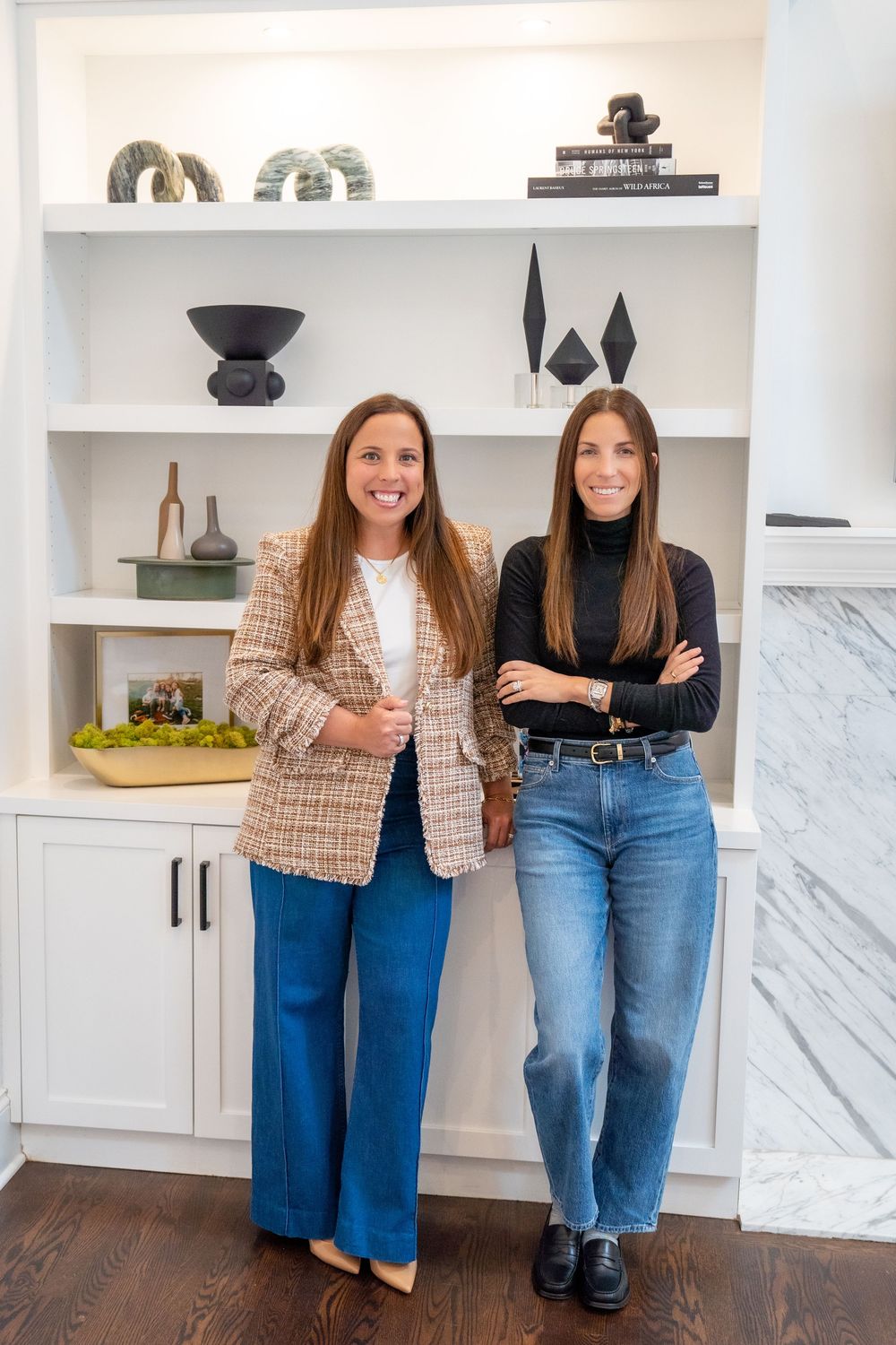 Two women smiling in front of white built-in shelves with decorative objects in a modern interior space.