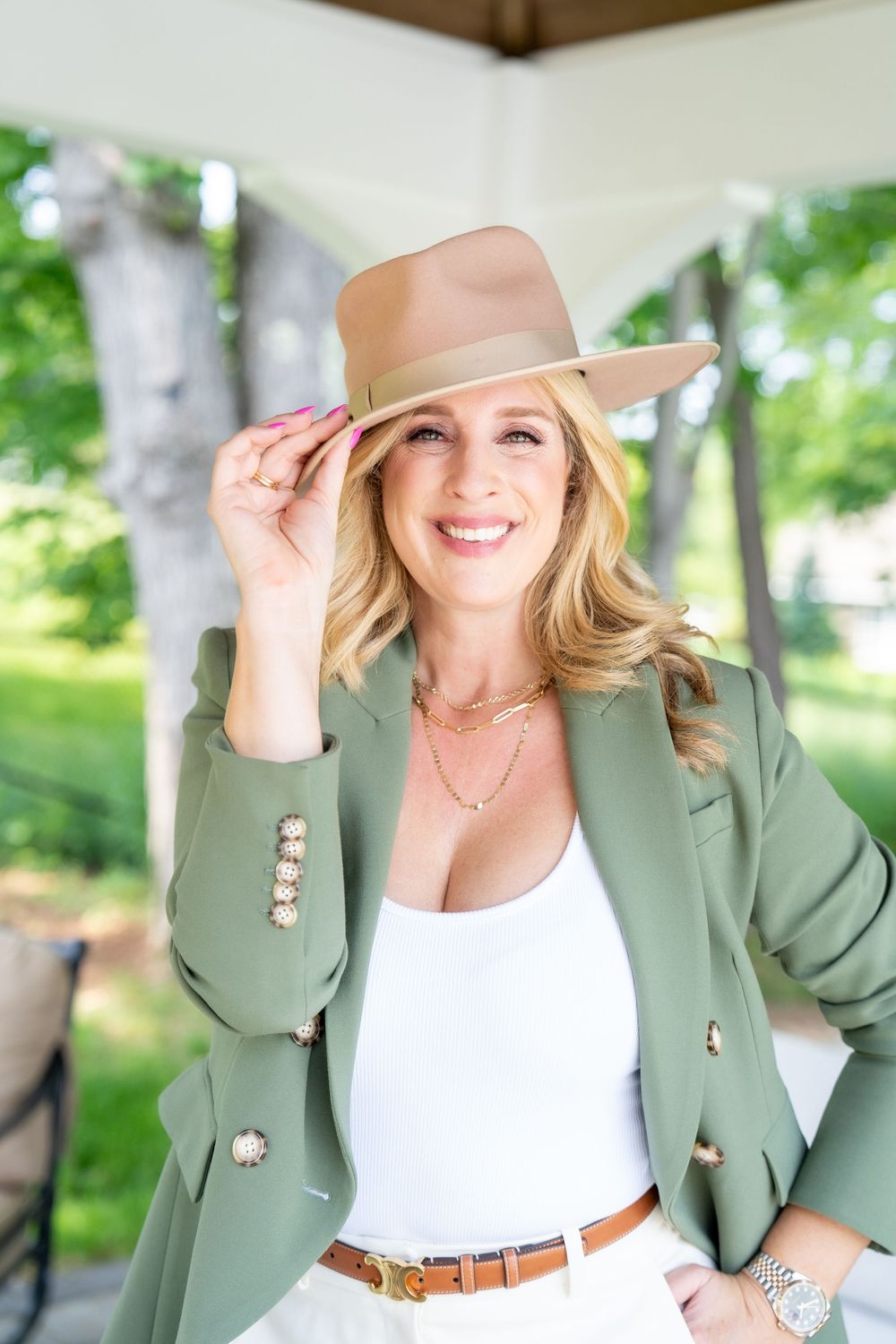 Smiling blonde woman in green blazer, white top, and tan fedora hat posing outdoors in a park setting.