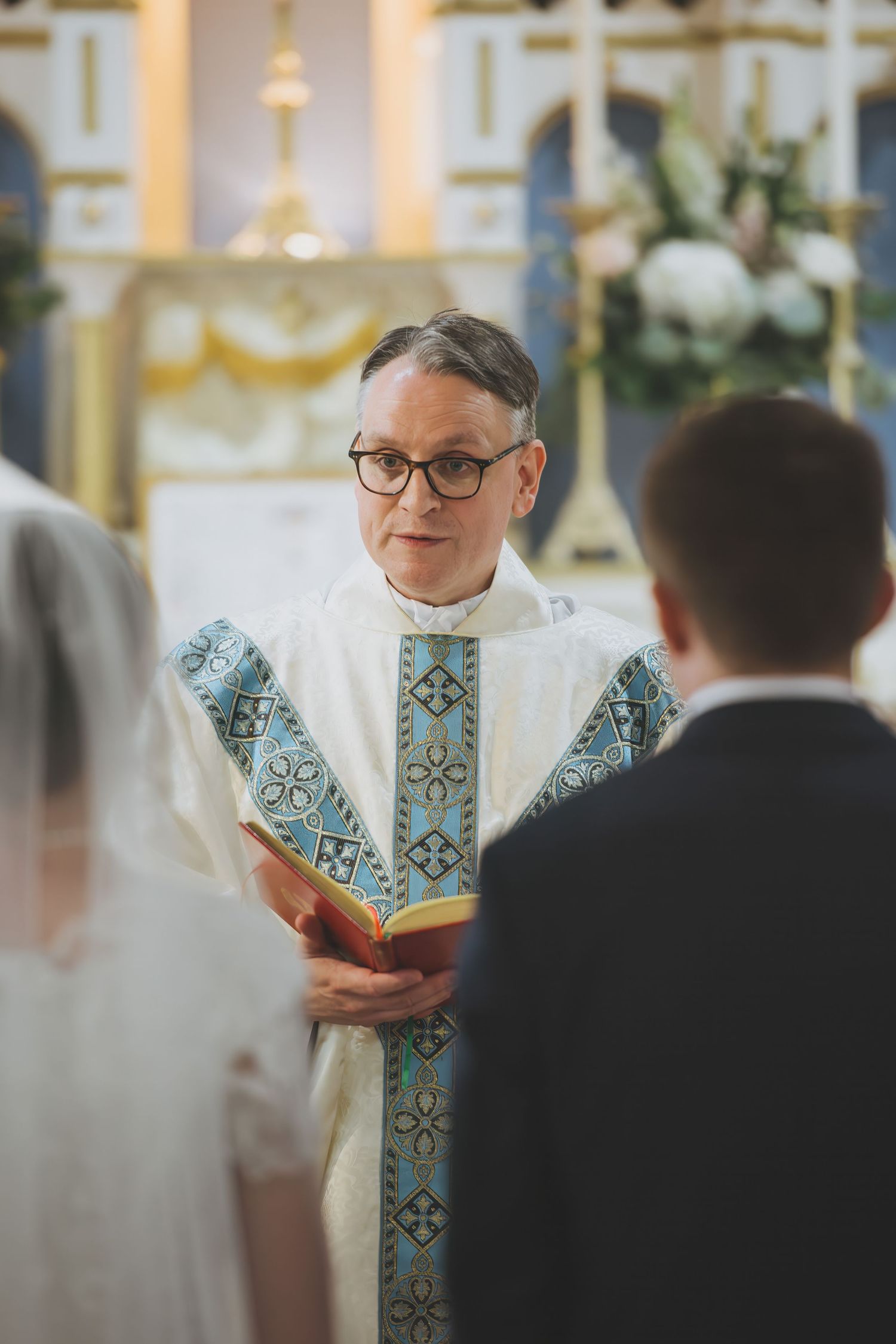 A priest in white and blue ornate vestments leads a wedding ceremony in an elegant church setting.