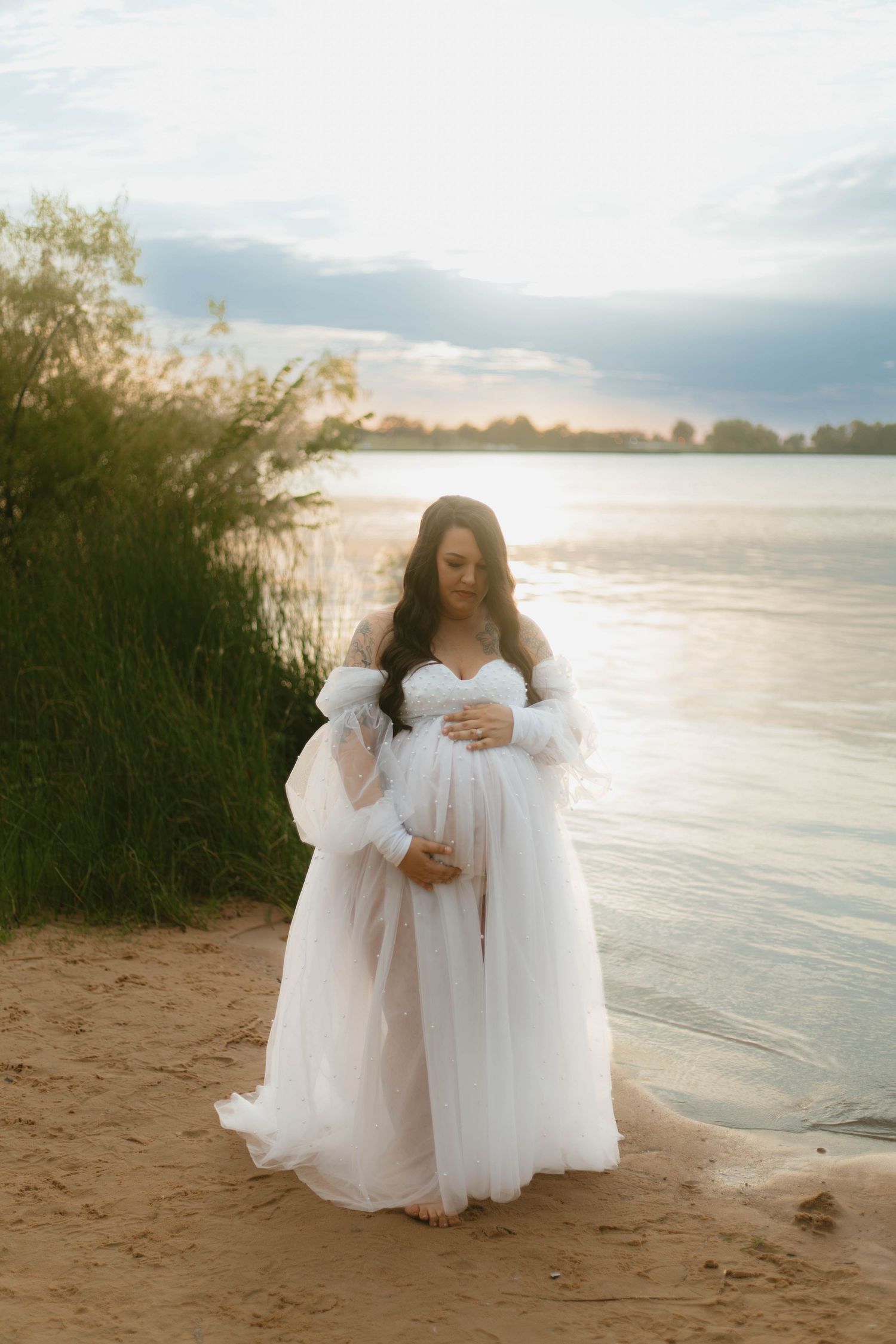 A maternity photo shoot on a sandy beach at sunset shows an ethereal white gown and glowing light over the water.