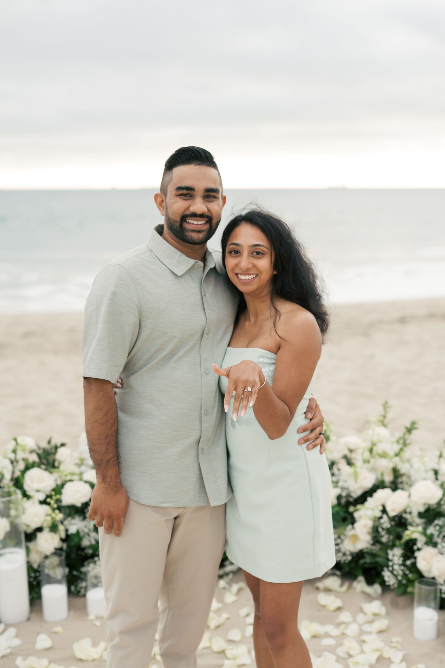 A romantic beach engagement photo shoot with white floral decorations and candles along the sandy shoreline at sunset.