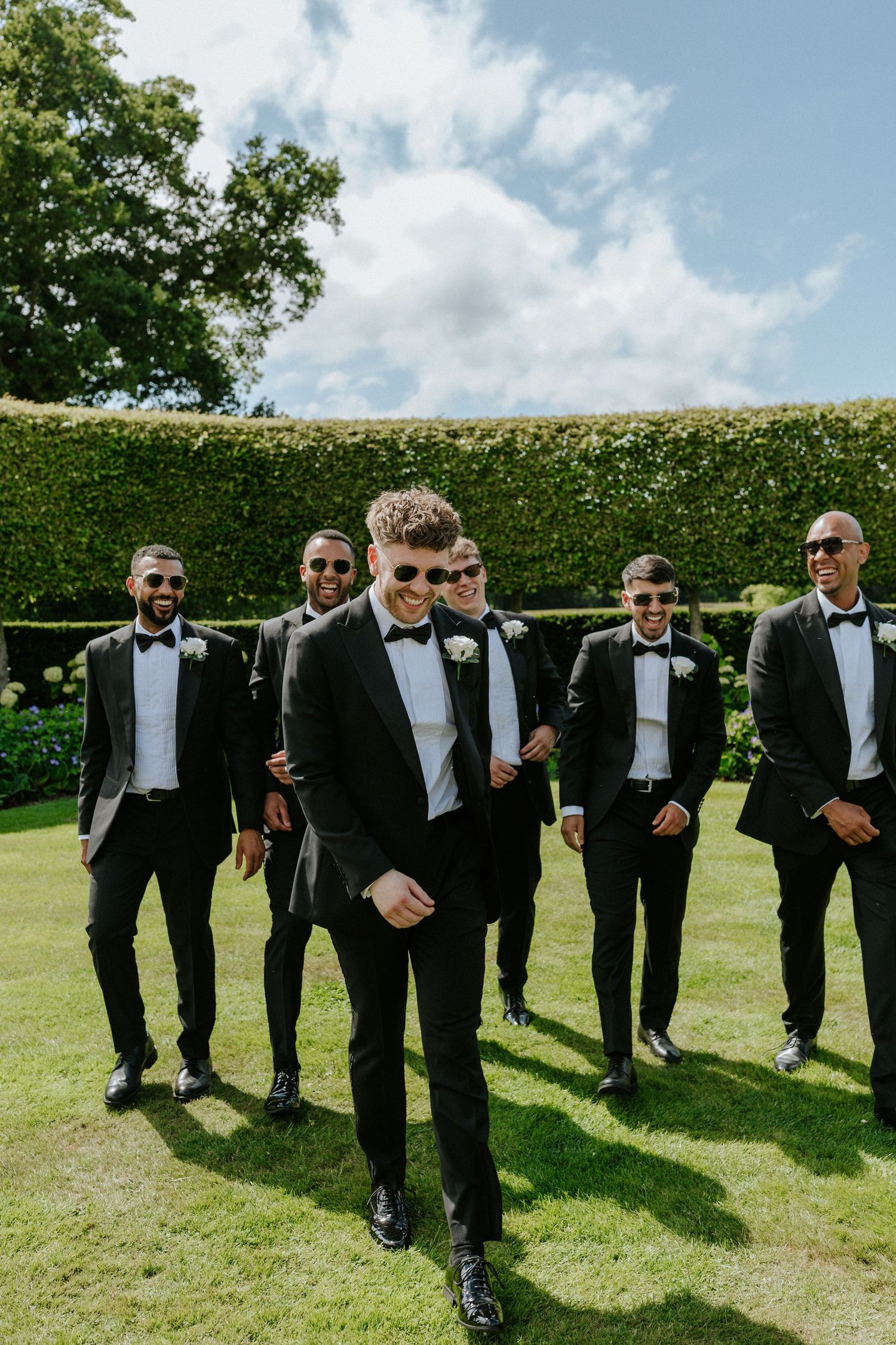 Groomsmen in black tuxedos and bowties pose for a formal photo on a manicured lawn with flowering garden backdrop.