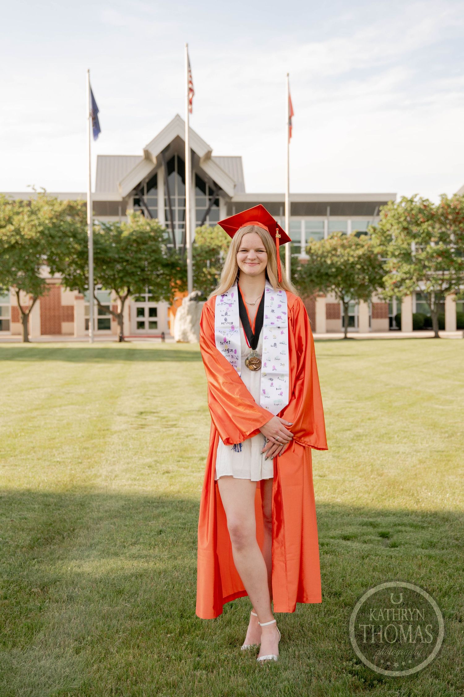 Graduate in orange cap and gown stands on campus lawn with academic building and flags in background.