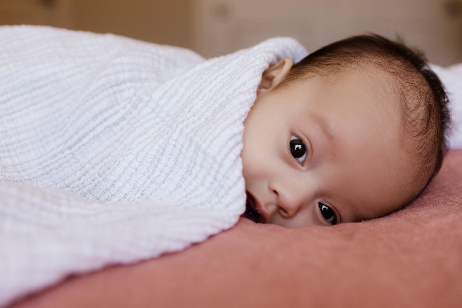 A small baby lies on a bed while looking towards the camera with wide eyes and a curious expression during in-home Bay Area session