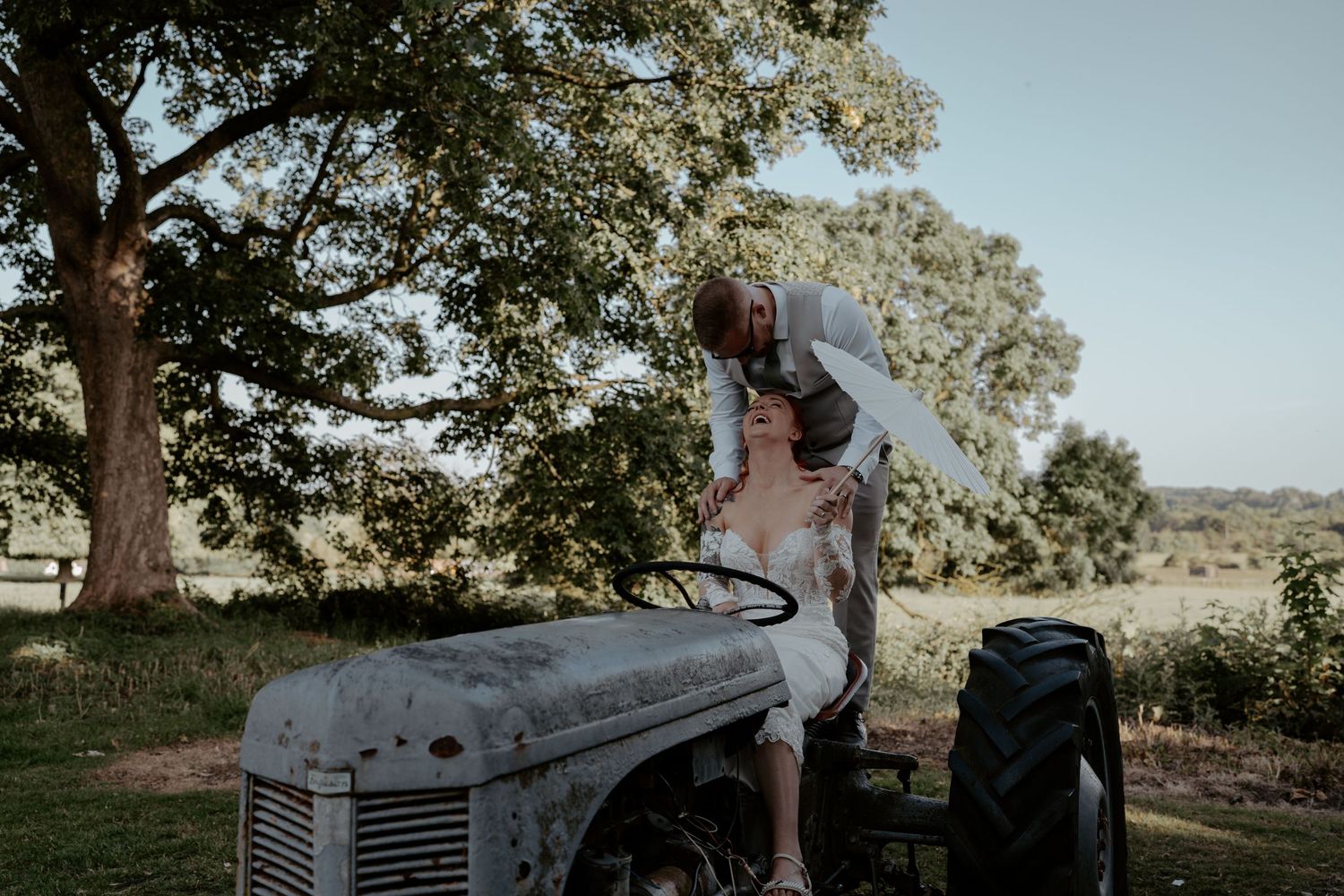 A couple stands in elegant wedding attire beneath a rustic brick archway with wooden doors at a historical venue.