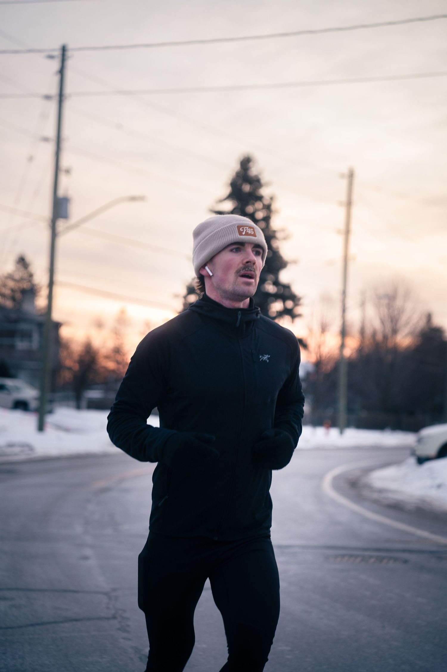 Person in a black winter coat and white beanie jogs on a snowy residential street at sunset.