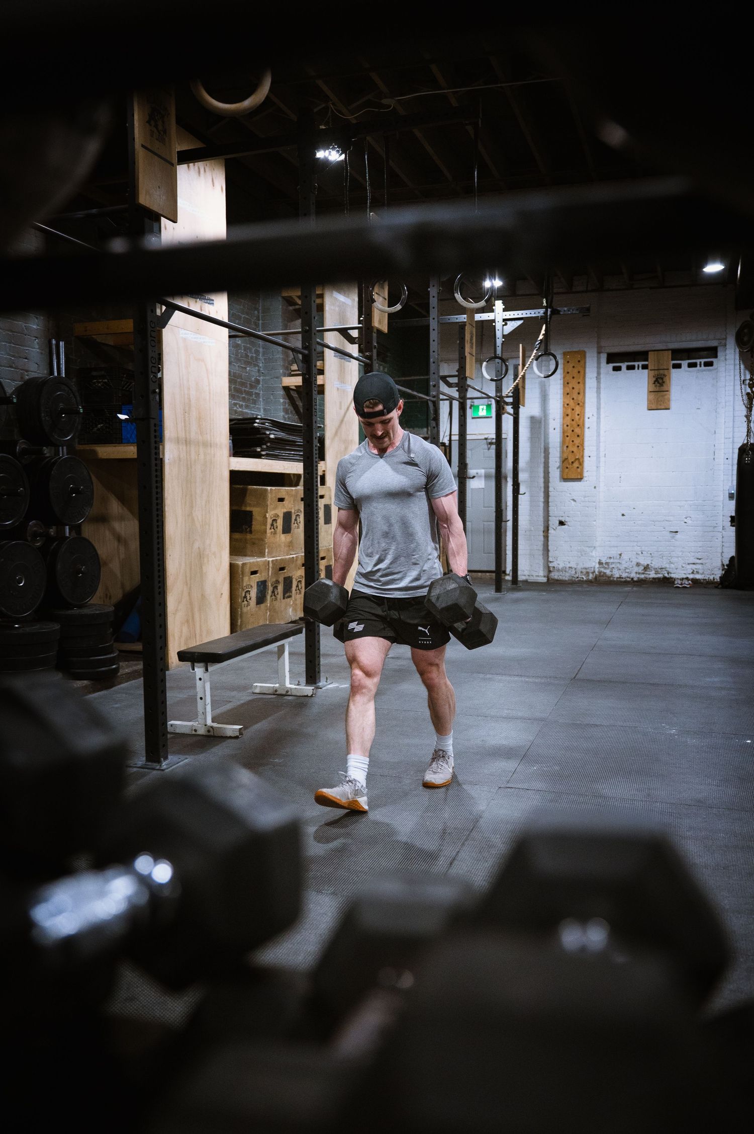 Athletic person working out in an industrial gym space with fitness equipment and dumbbells visible in the dark setting.