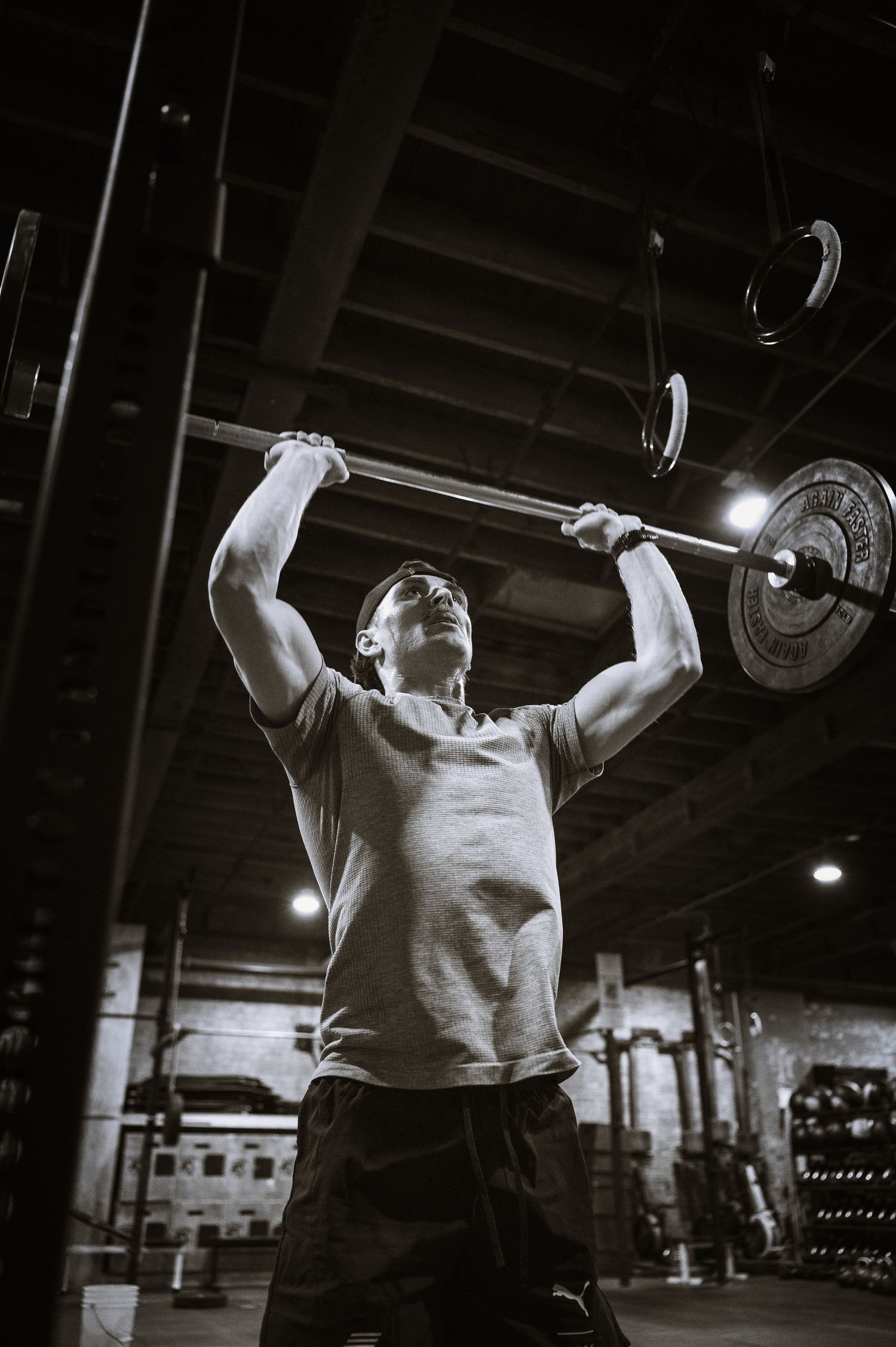 Black and white photo of an athlete performing an overhead barbell press during an intense workout at a gym.