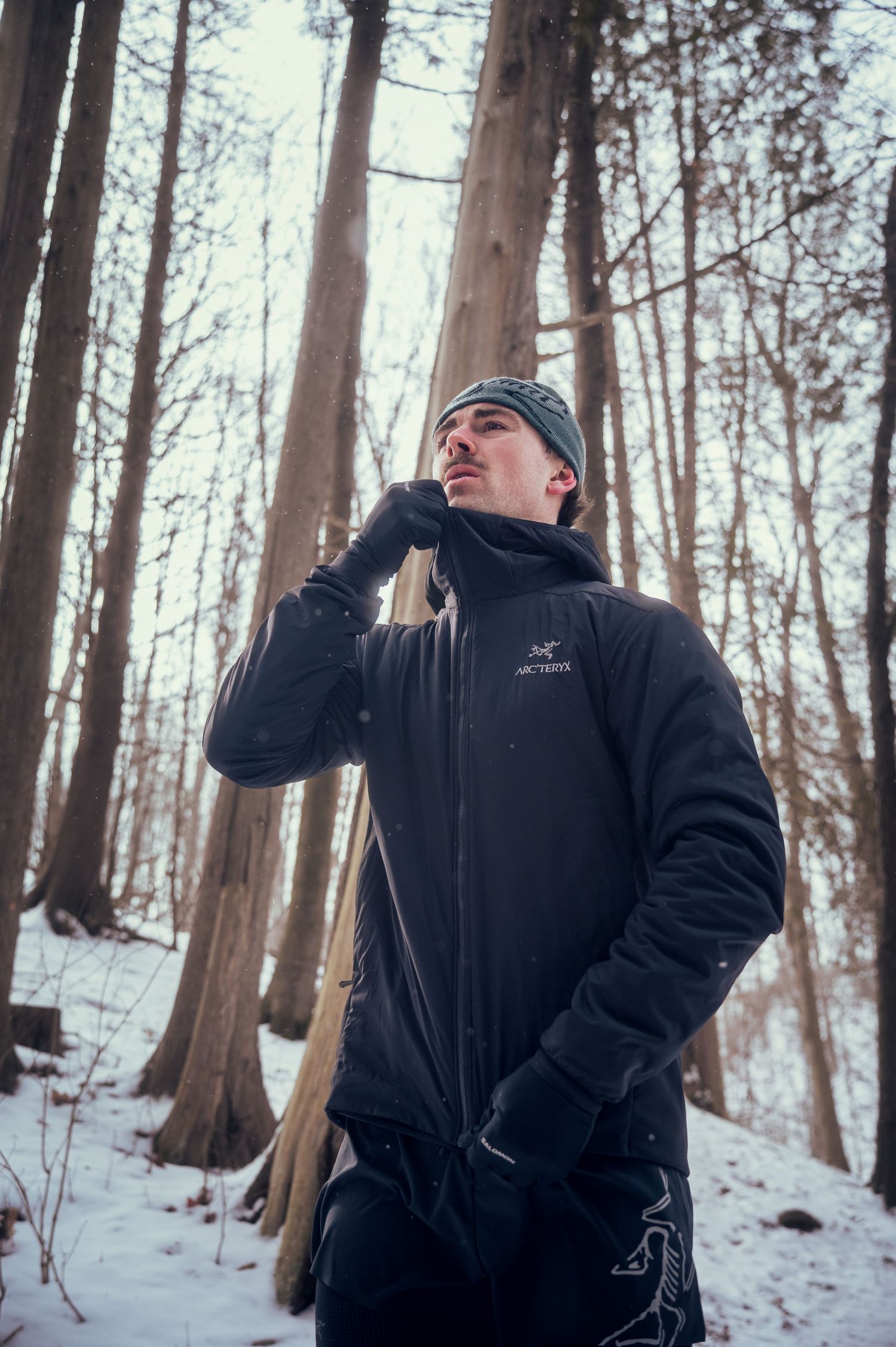 Person in winter clothing poses playfully in a snowy forest setting with bare trees in the background.