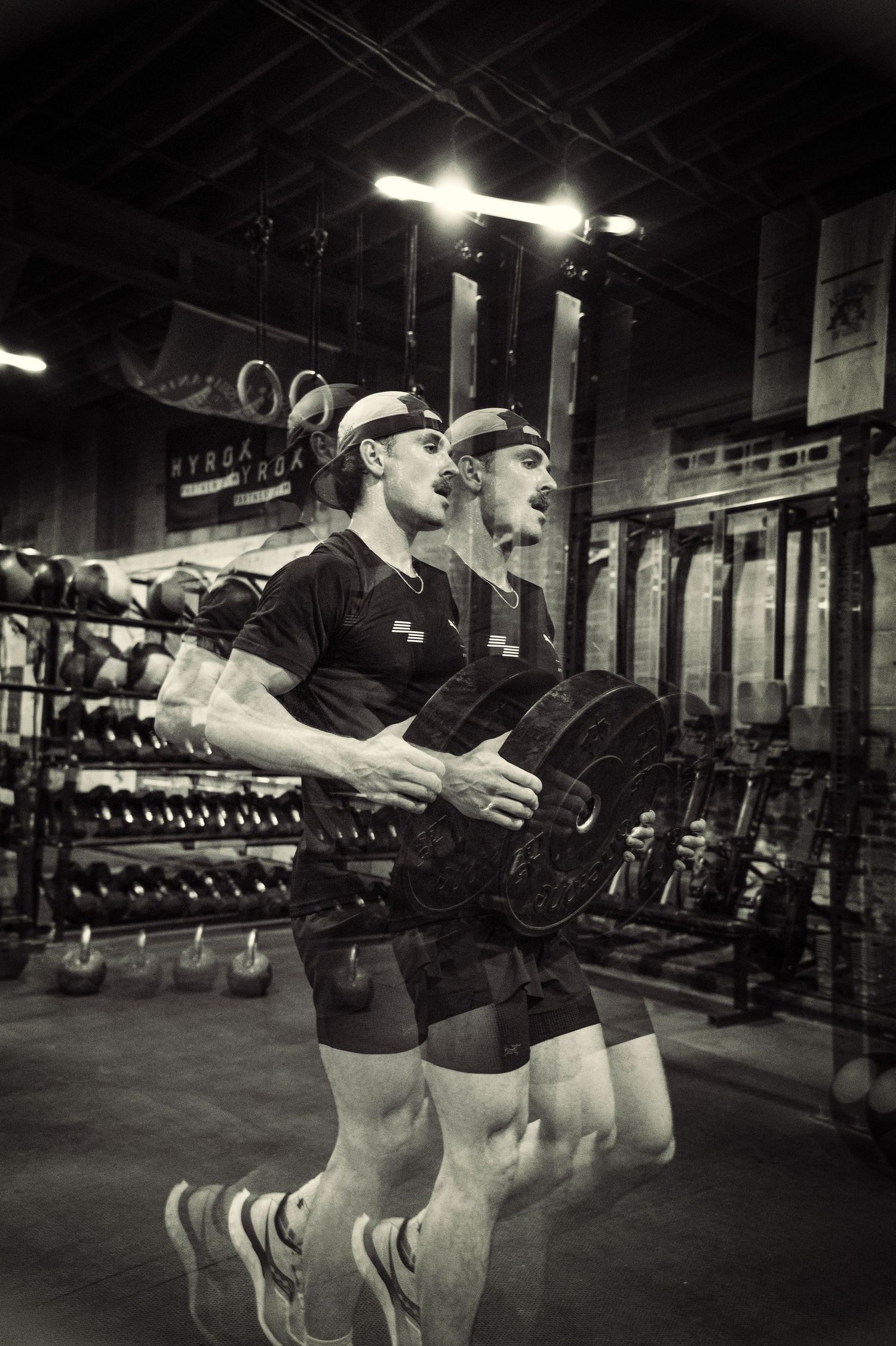 Black and white photo of athletes performing synchronized barbell exercises in a gym with overhead lighting.