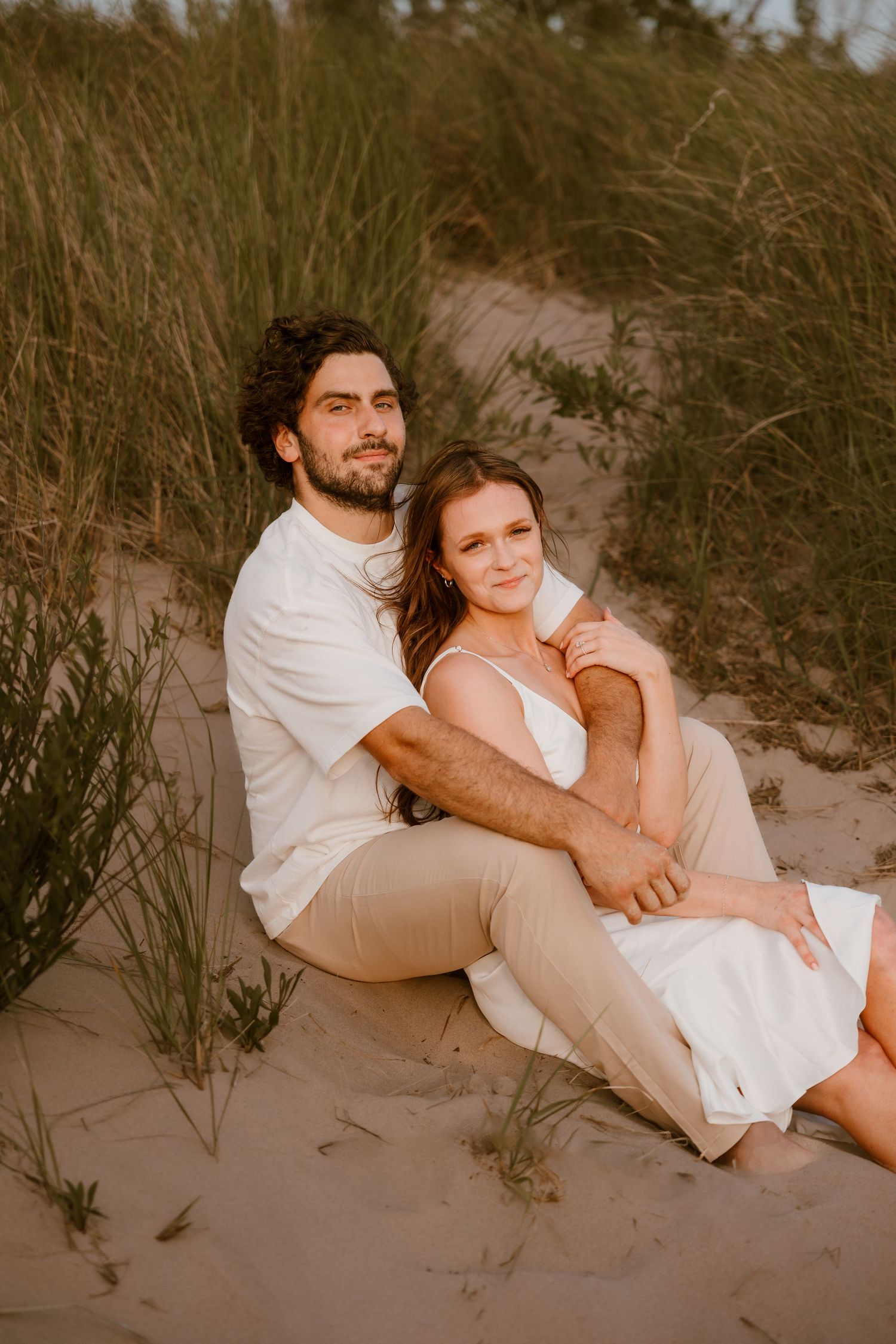Romantic couple in white outfits embracing on sandy beach dunes during golden hour photoshoot series. Michigan Engagement Session