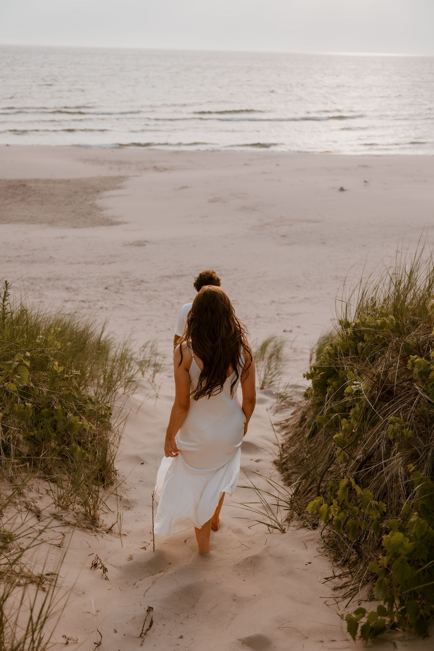 A figure in a flowing white dress walks down a sandy beach path through sea grass at sunset in a dreamy scene. Detroit Wedding Photographer