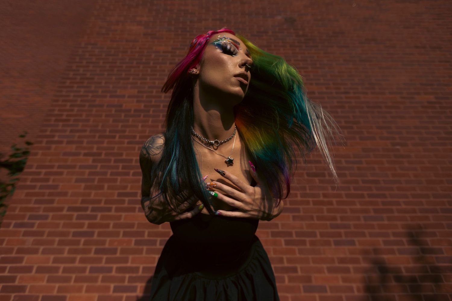 Alternative fashion model with vibrant rainbow hair poses dramatically against brick wall wearing sunglasses.