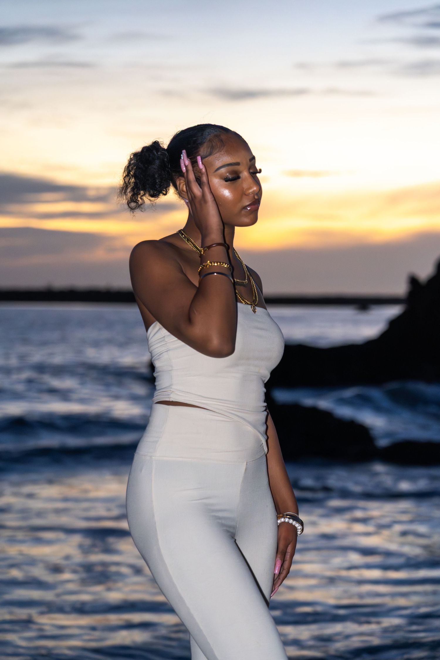 Woman in white outfit poses against sunset ocean backdrop with hands near face.