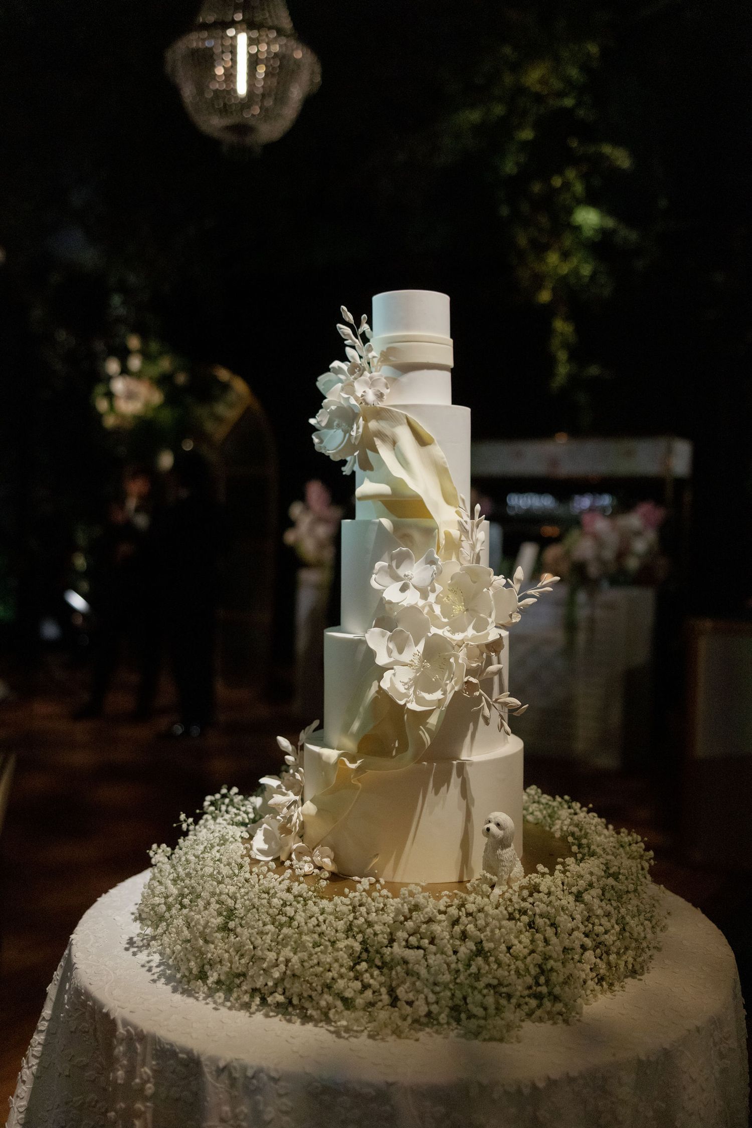 Elegant three-tiered white wedding cake decorated with delicate flowers and baby's breath on a dark background.