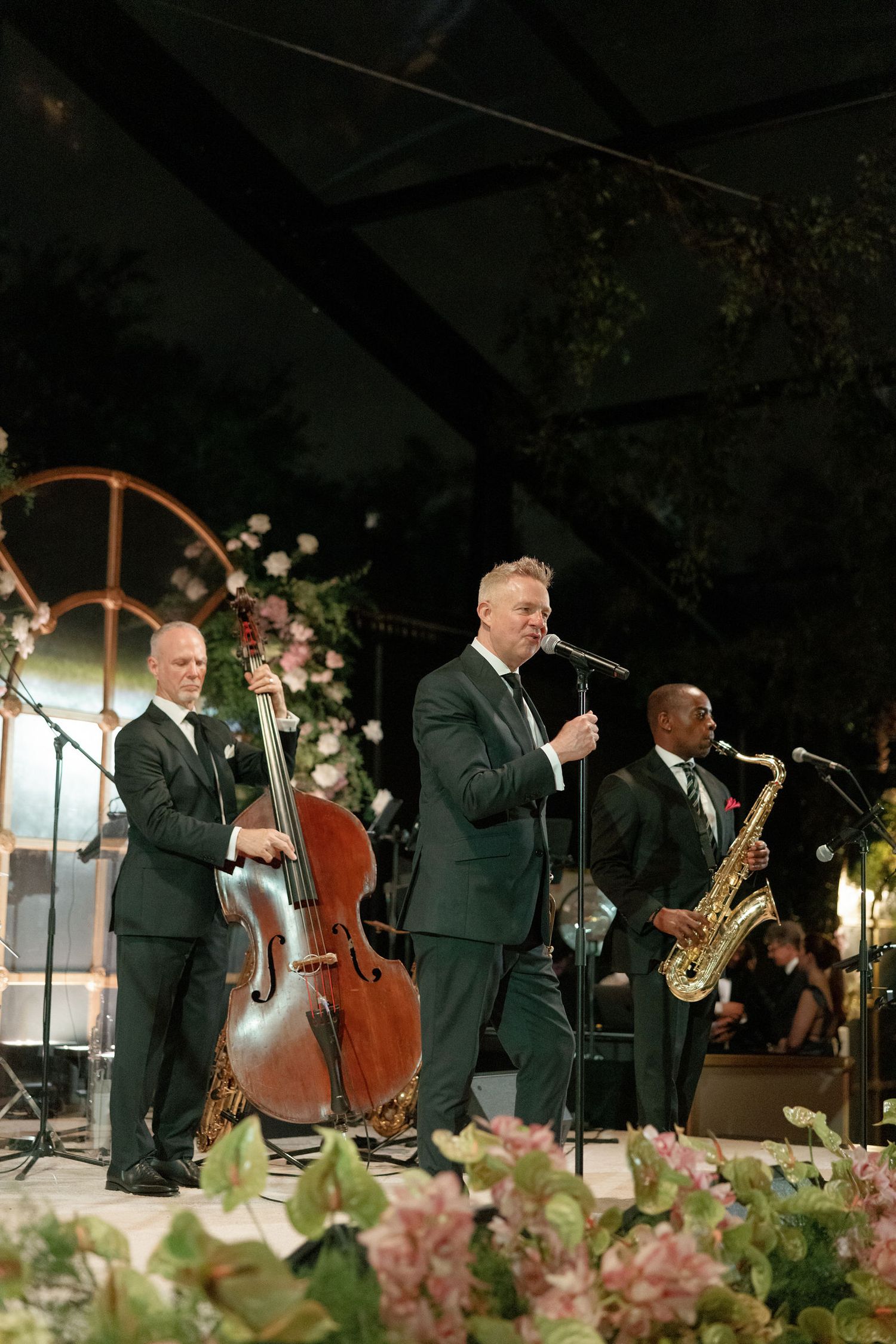 A jazz band performs at an outdoor event with floral decorations, featuring a bassist, singer and saxophone player.