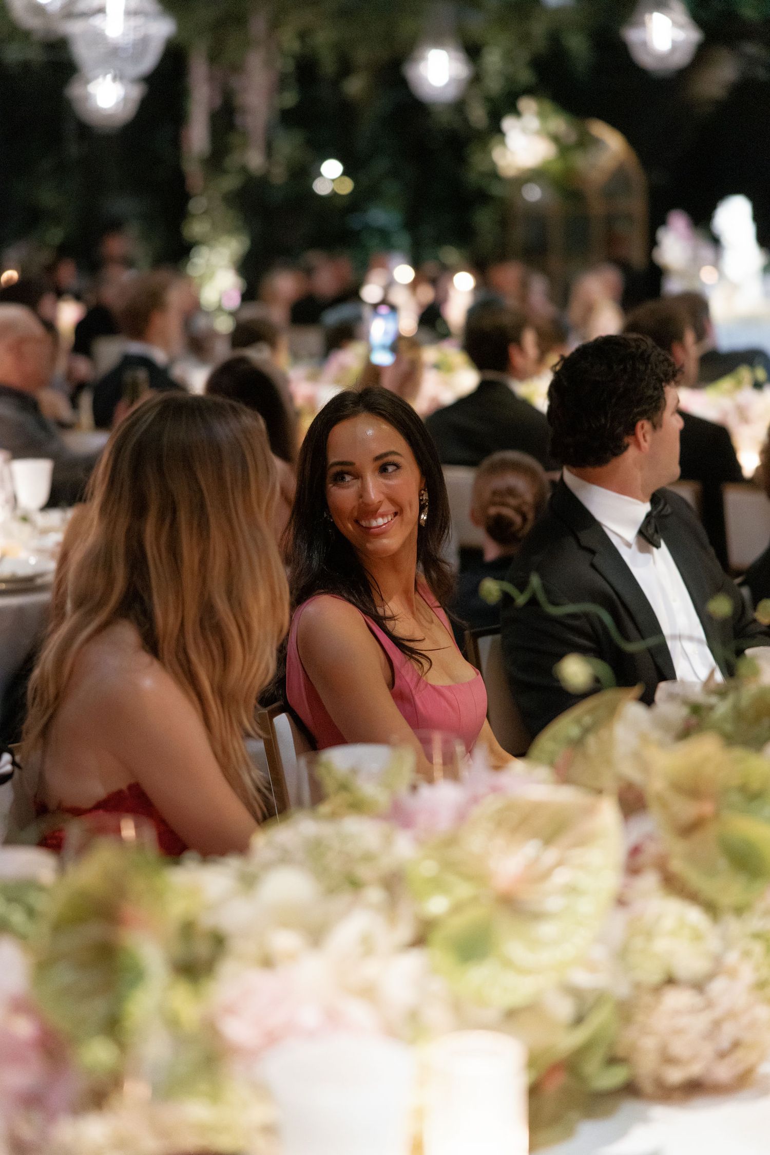 Wedding guests in formal attire engage in conversation at an elegantly decorated dinner table with floral centerpieces.