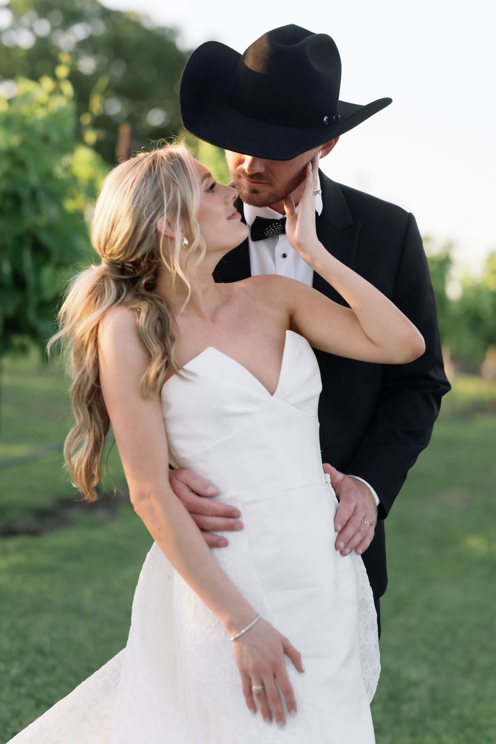 A romantic wedding couple embraces in an outdoor garden setting while wearing western-themed formal attire.