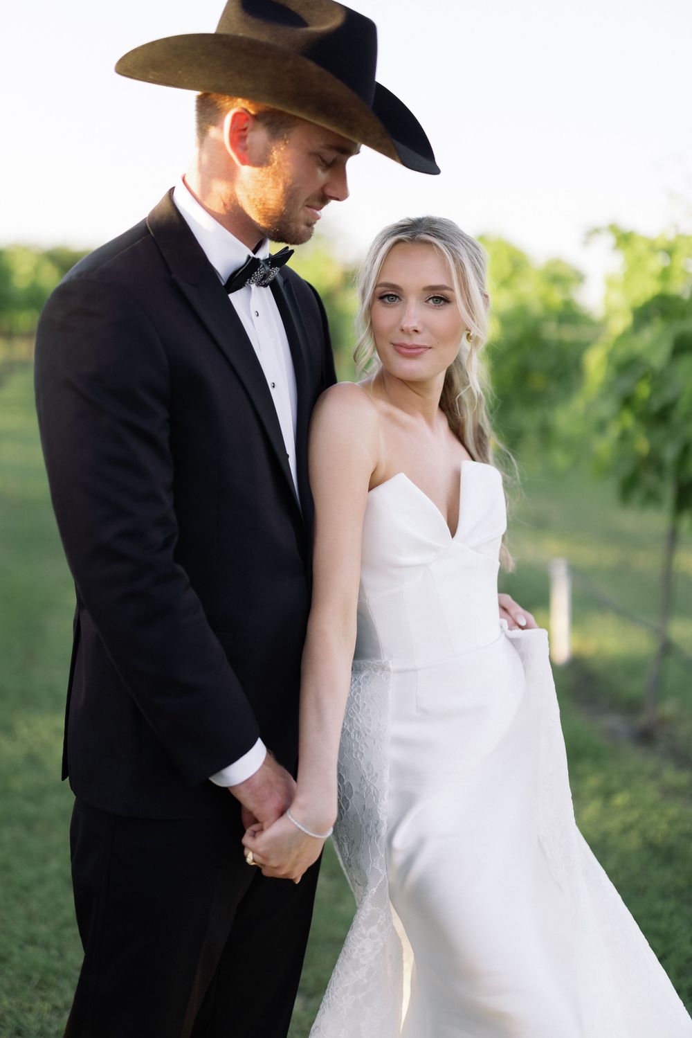 Stylish wedding couple in western-inspired formal wear pose together in vineyard setting.