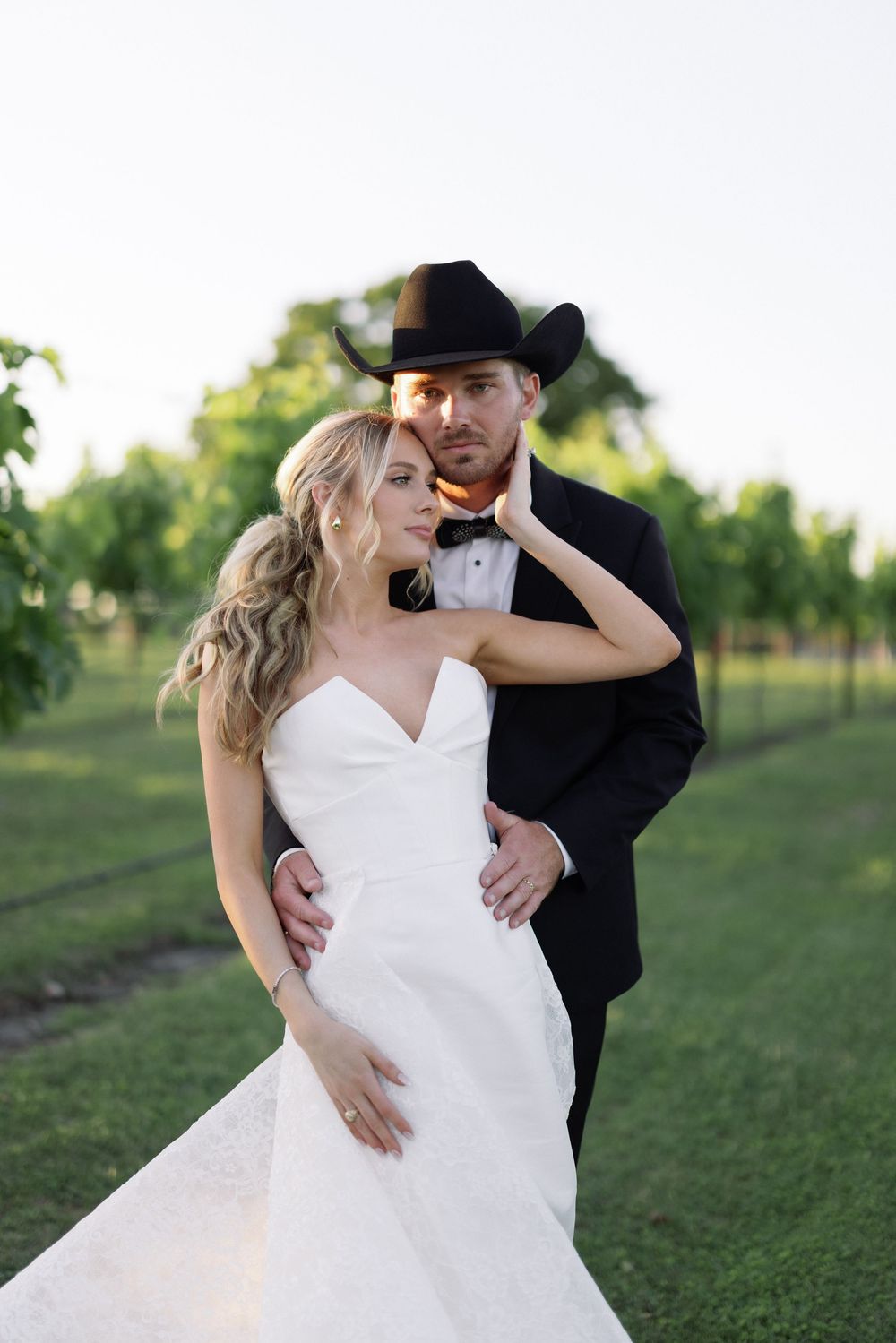 Elegant couple in wedding attire pose in a vineyard with groom wearing a western style black cowboy hat.