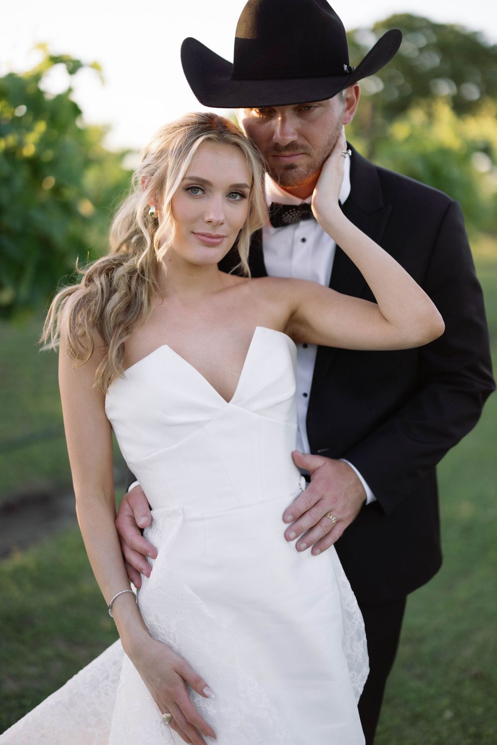 Romantic wedding portrait featuring western style with cowboy hat and elegant white bridal gown in outdoor setting.