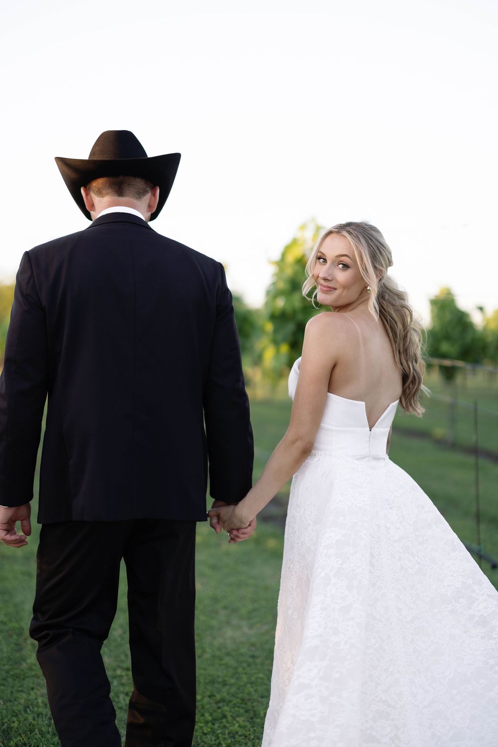 A couple in formal wedding attire walks through a grassy field on their special day.
